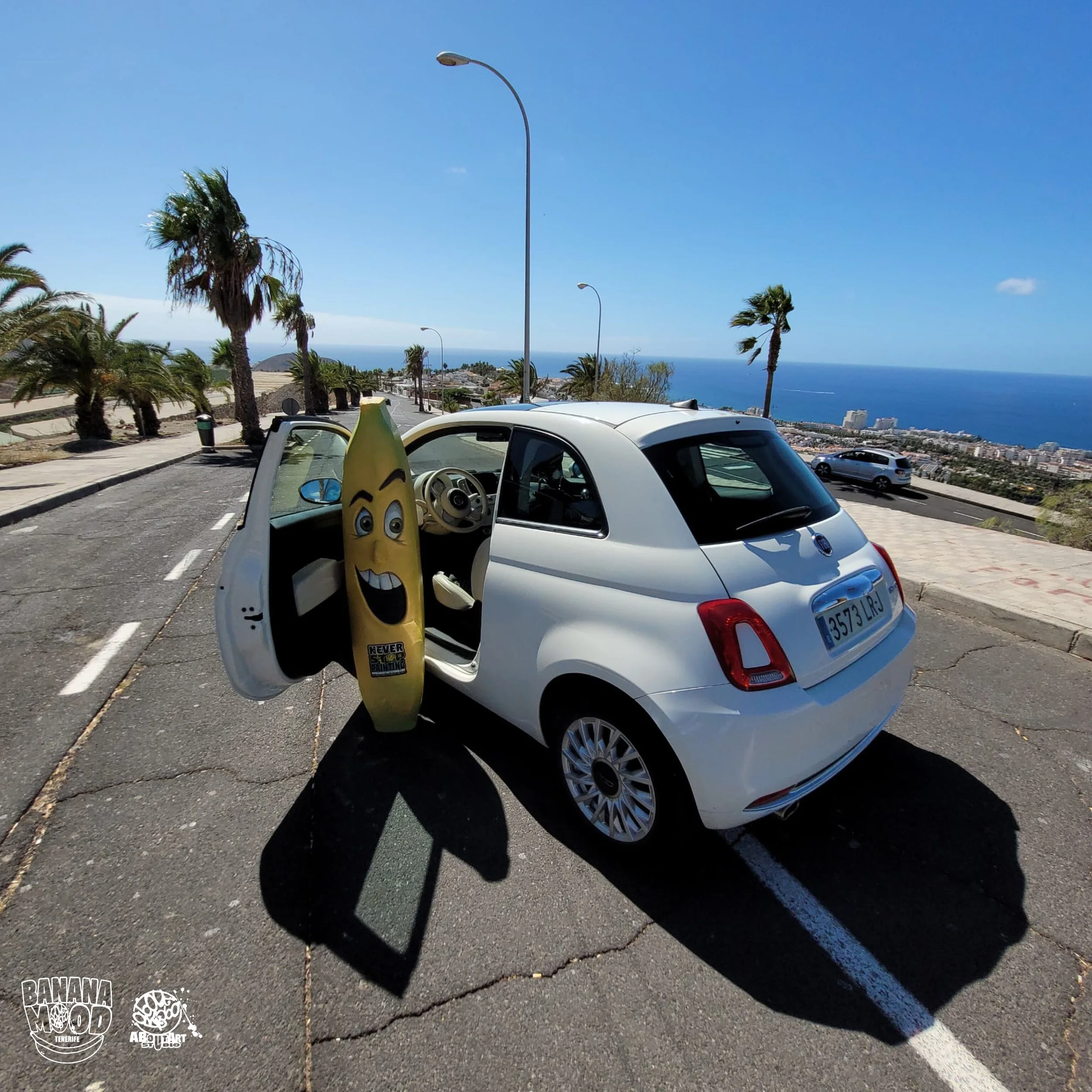 Auto blanco de dos puertas en un mirador con vista al mar, con una banana de peluche en la puerta abierta, palmeras y un cielo despejado.