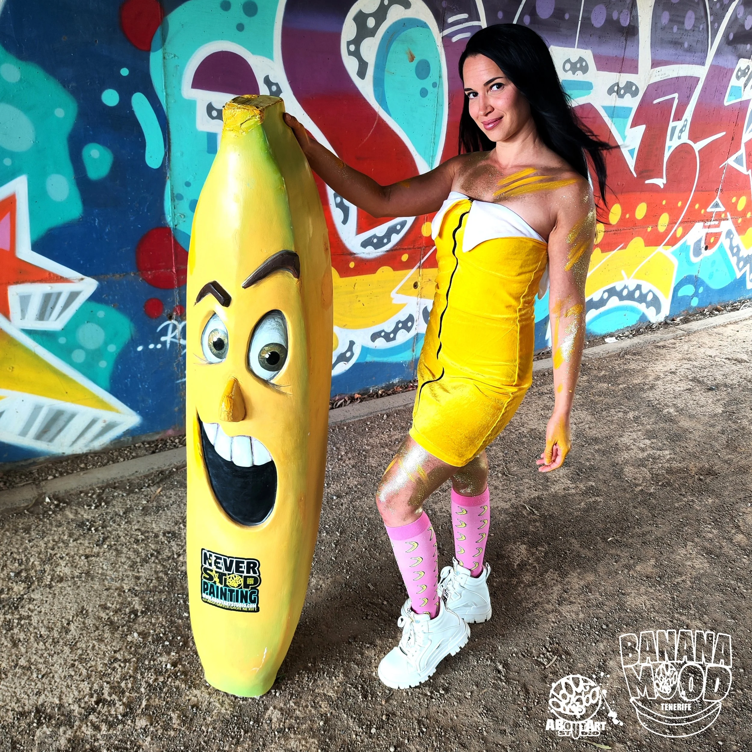 Una mujer con ropa amarilla y accesorios rosas, pintada con spray dorado, posa junto a un cartel de banana con cara alegre en un fondo de graffiti colorido.