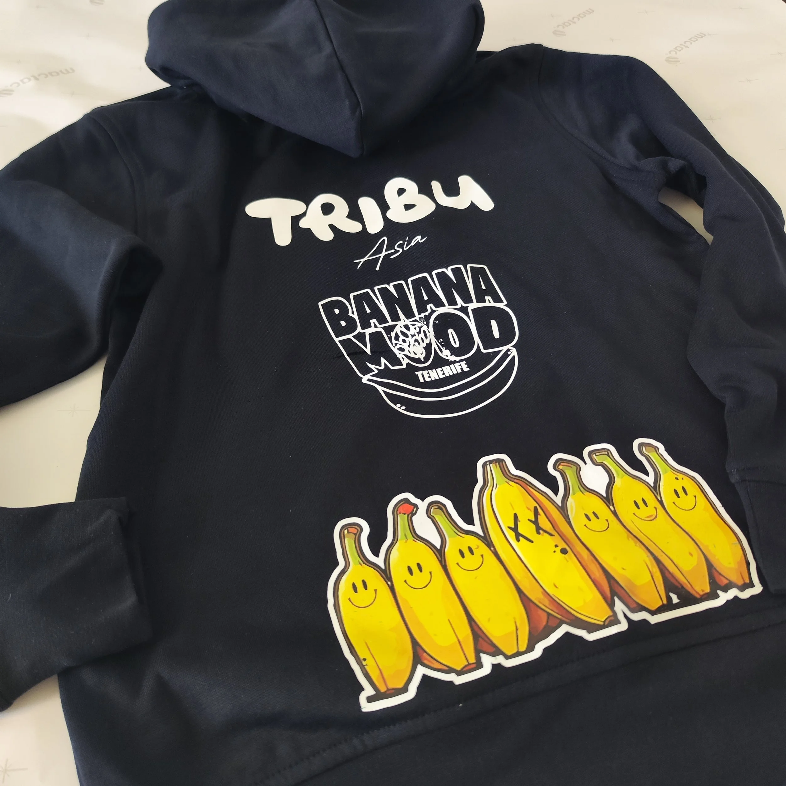 Sudadera negra con logotipo de la marca 'Tribu Asia', la palabra 'Banana Mood' con una ilustración de plátanos, algunas con caras sonrientes y otras con caras de 'X' en los ojos, y la palabra 'Tenerife' debajo.