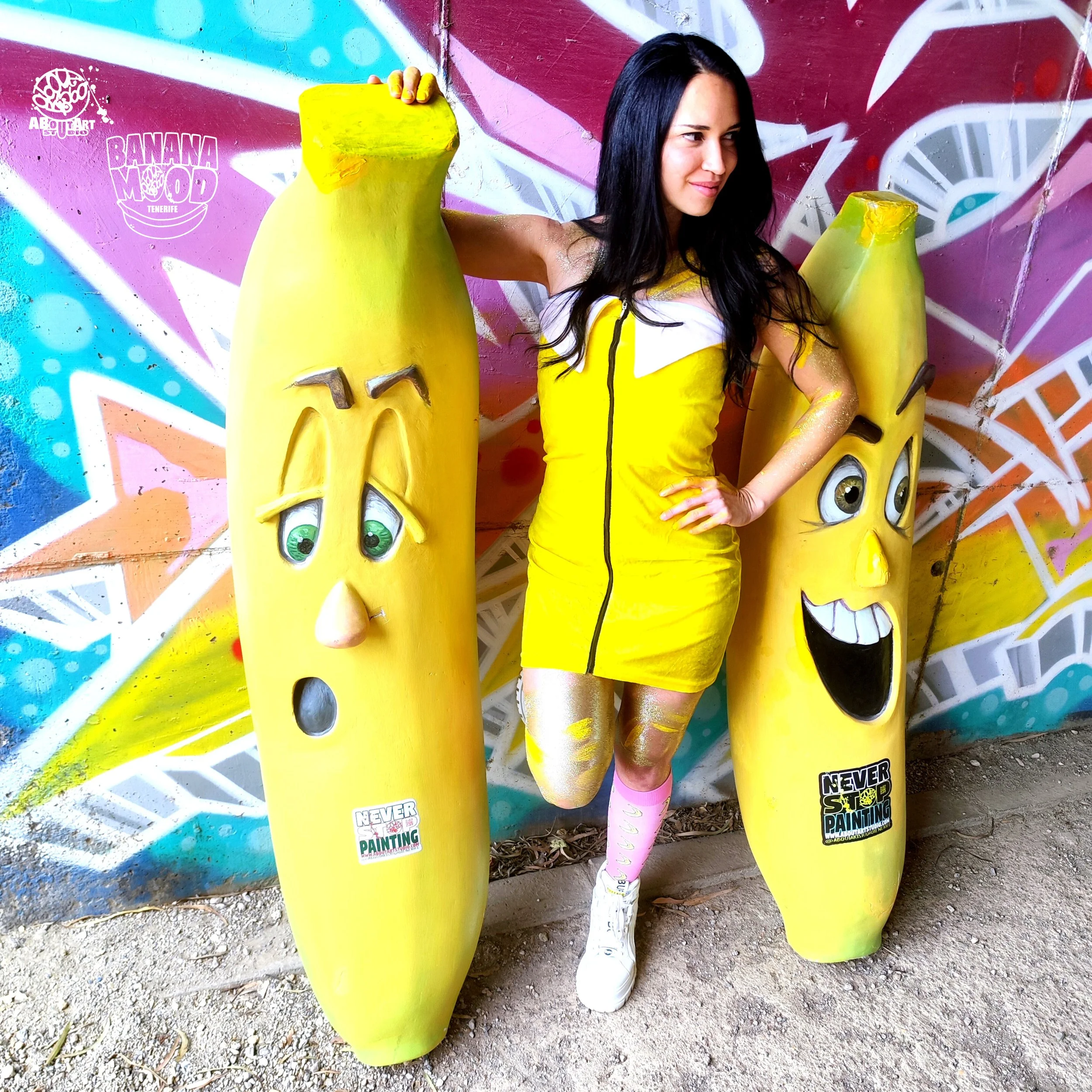 Una mujer con vestido amarillo y calcetas rosadas posa entre dos estatuas de plátanos con rostros expresivos, en un fondo de arte callejero colorido.