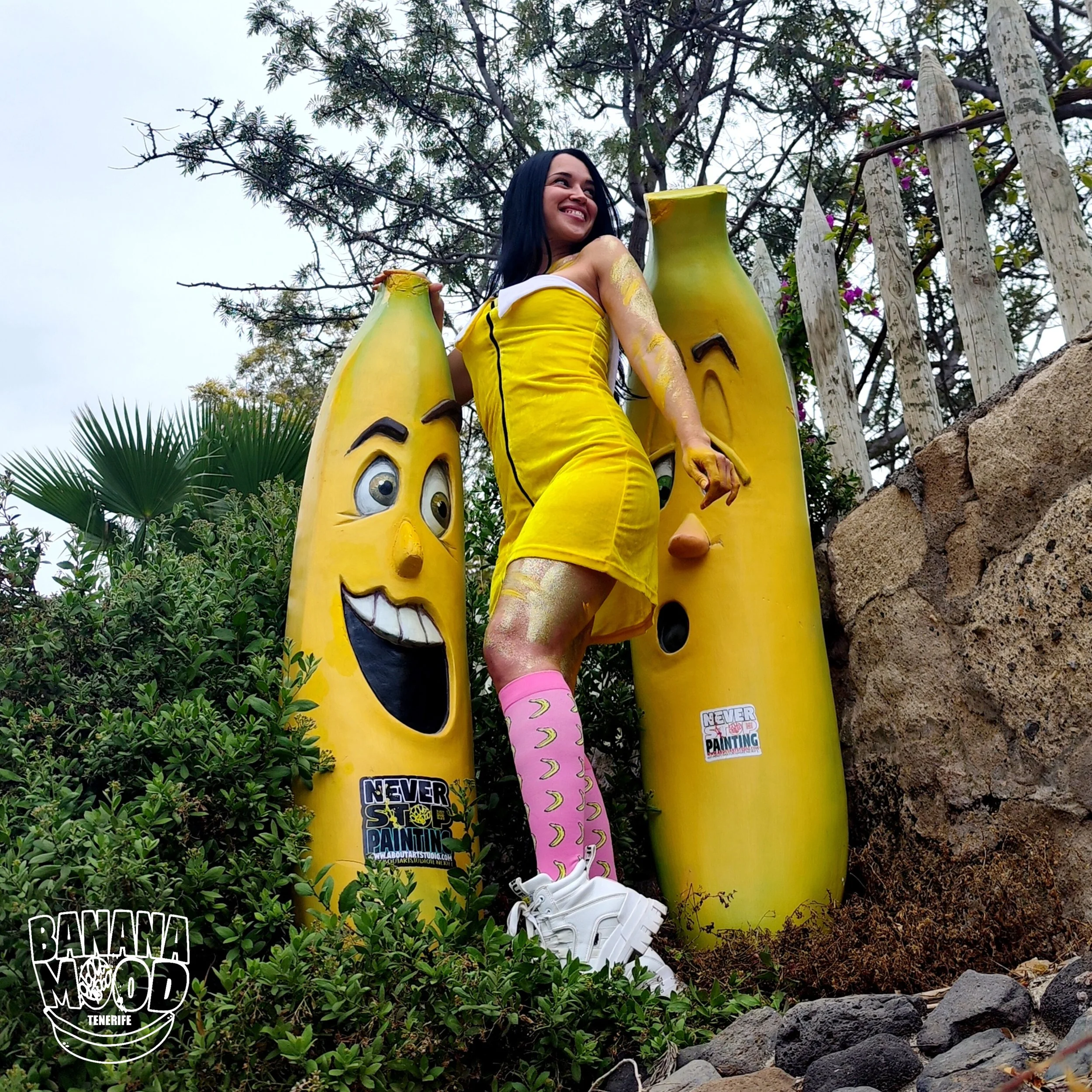 Una mujer de vestido amarillo con medias rosadas y zapatillas blancas está posando entre estatuas de plátanos con caras sonrientes en un entorno al aire libre con vegetación y un cielo nublado.