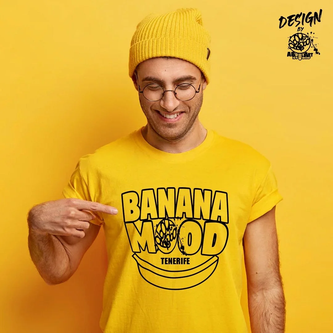 Hombre con camiseta amarilla y gorro amarillo señalando una camiseta similar que dice "Banana Mood Tenerife" con dibujo de banana y fondo amarillo.