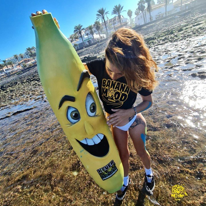 Una mujer con cabello rizado rubio, usando camiseta negra con letras amarillas que dicen 'BANANA MOOD', posa con una tabla de surf con cara de plátano en la playa, con palmeras y casas blancas en el fondo. La tabla tiene una expresión facial divertid