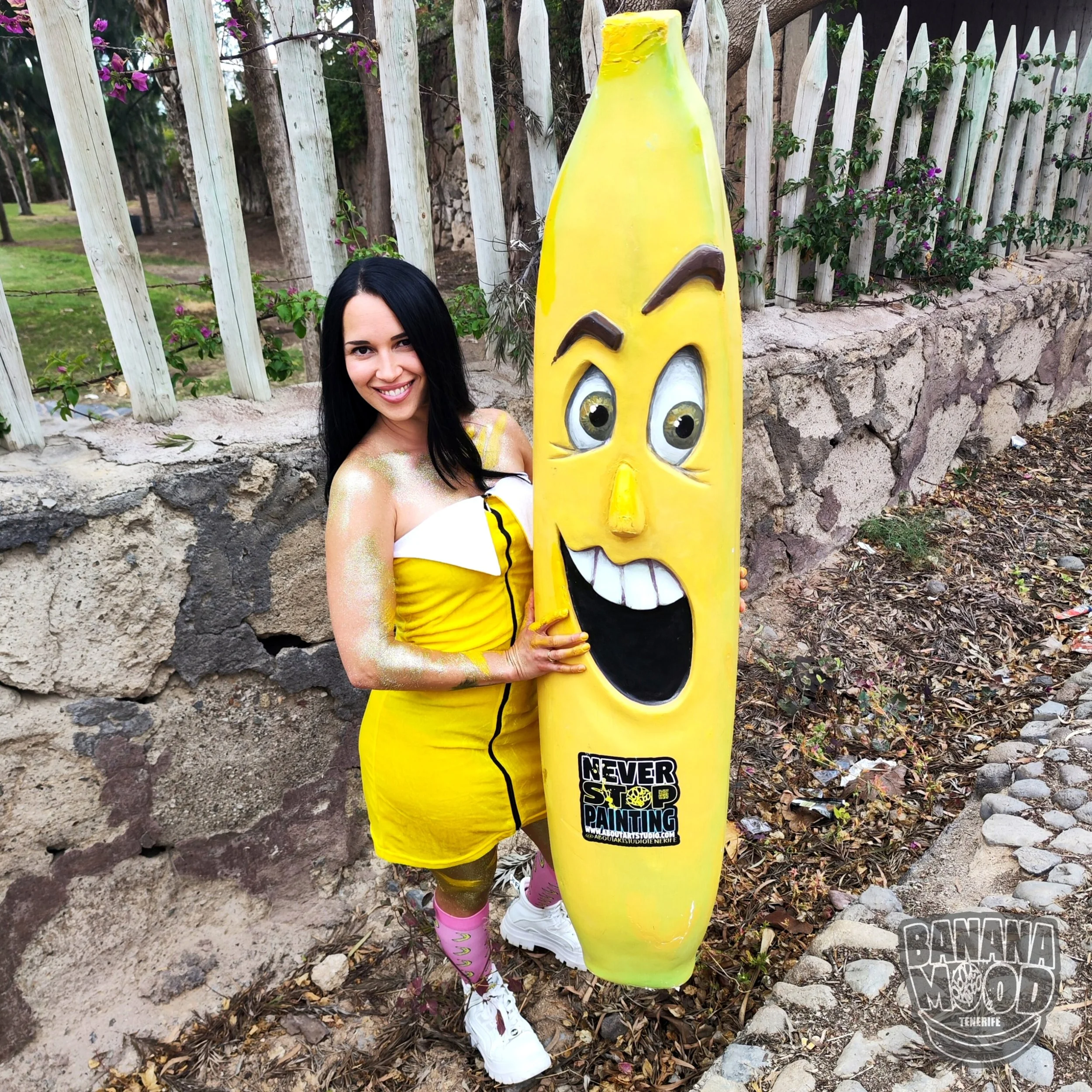 Una mujer con vestido amarillo y medias rosas posando junto a una figura de arte callejero en forma de plátano con cara feliz en un entorno al aire libre con cerca de madera y vegetación.