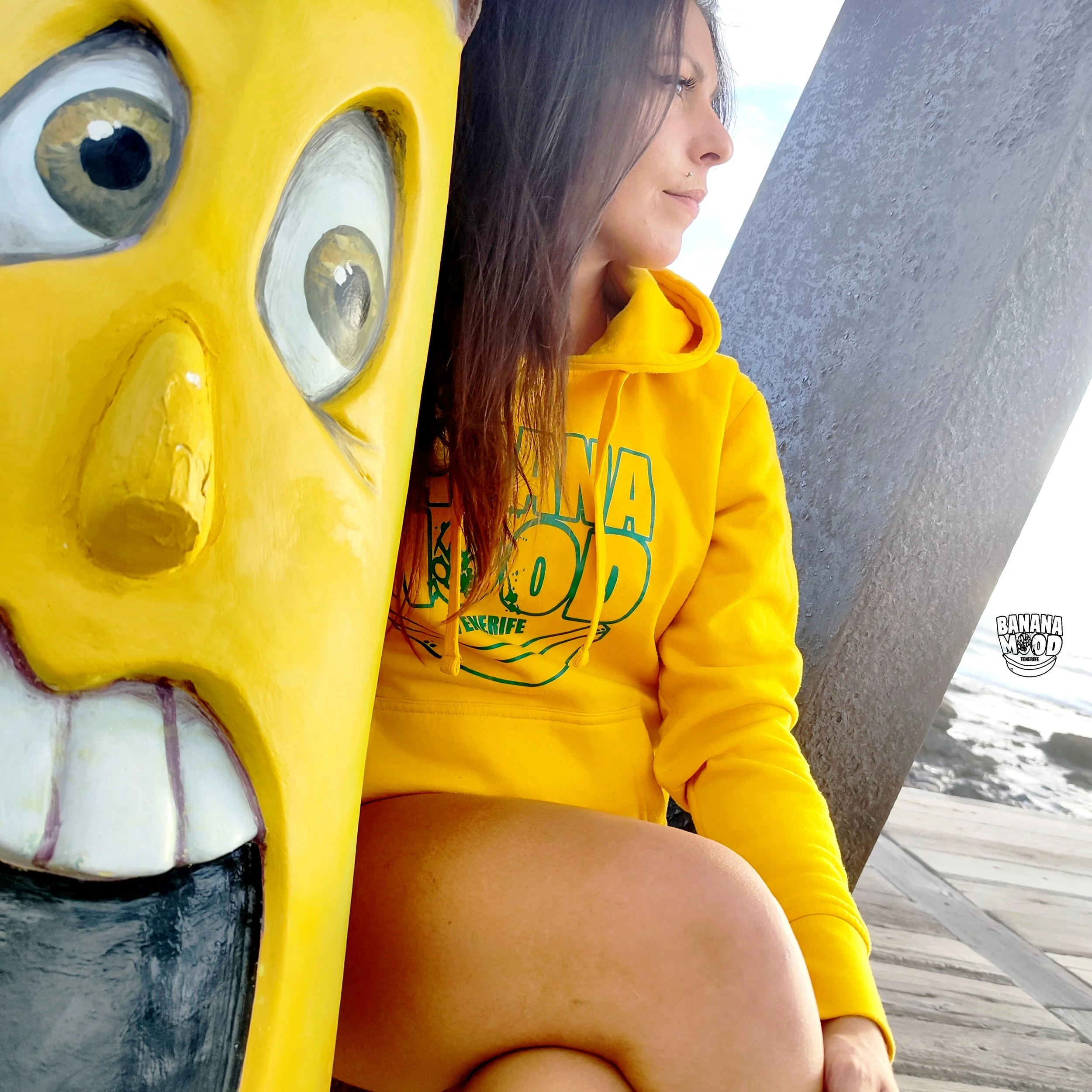 Joven mujer con sudadera amarilla, sentada junto a una escultura amarilla con rostro, en un lugar exterior junto a una estructura de metal y camino de madera.