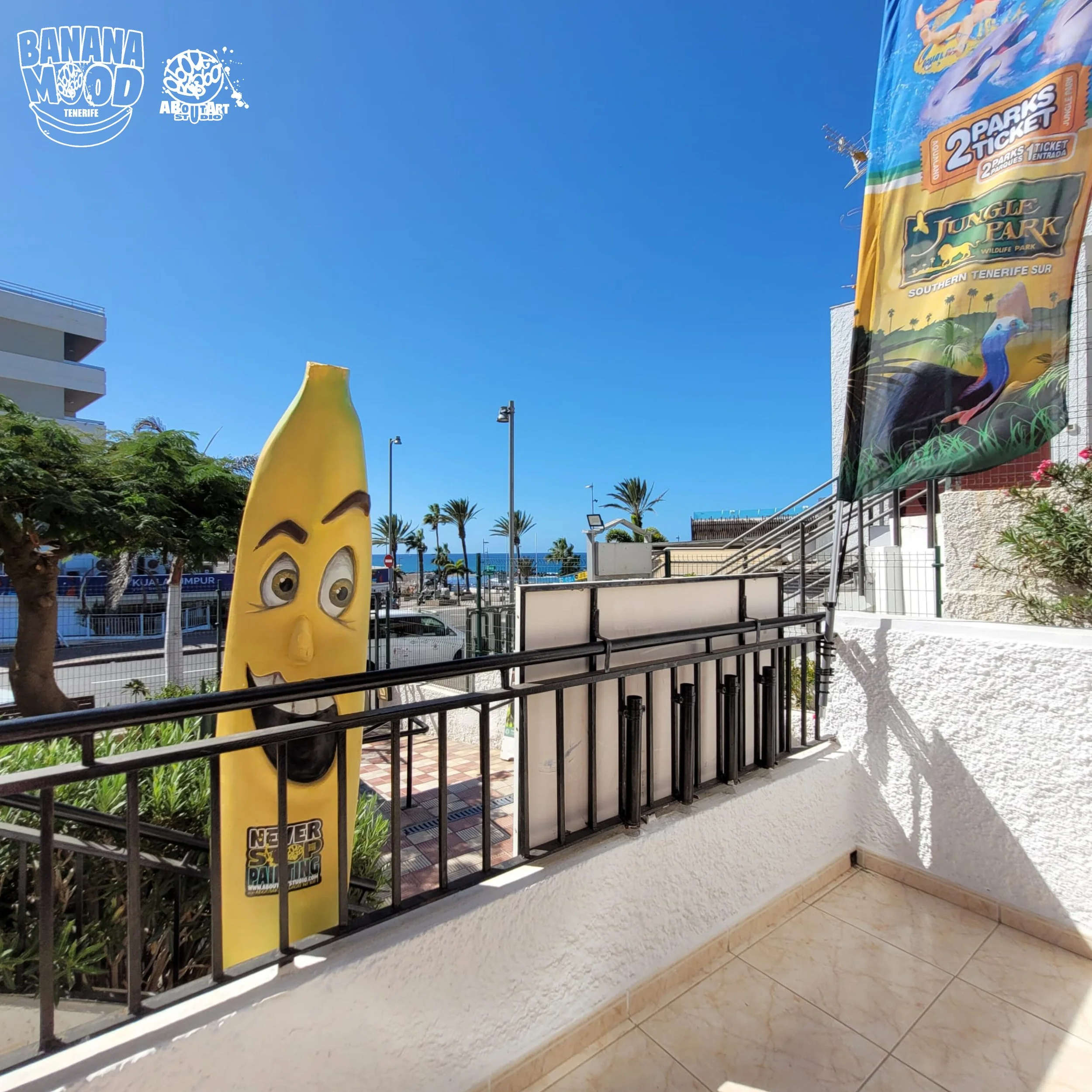 Una figura de un plátano con rostro sonriente y ojos grandes, ubicada en un balcón con vallas negras y presencia de una bandera promocional del parque Jungle Park en Tenerife, con palmeras y un cielo despejado de fondo.