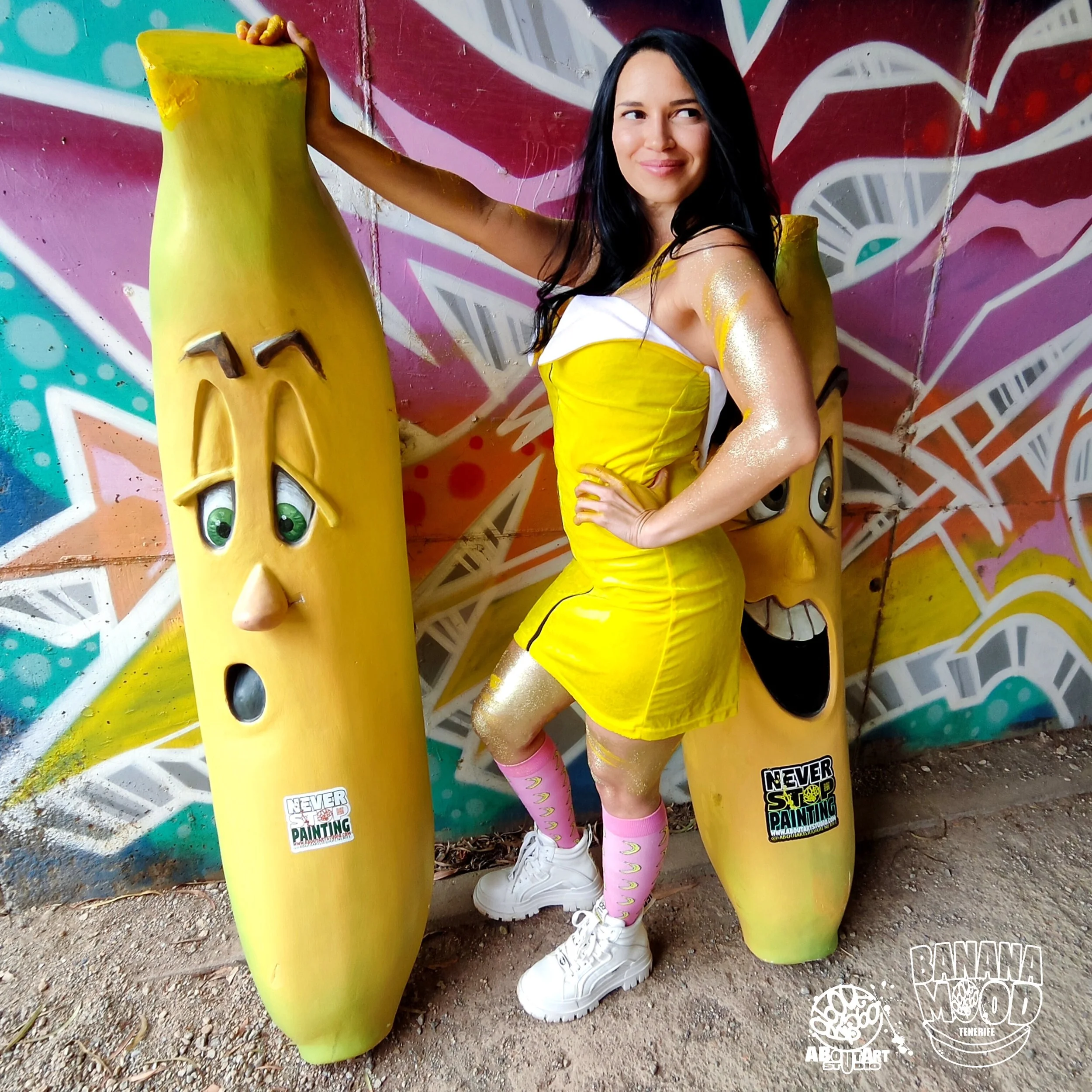 Mujer con ropa amarilla y camiseta blanca posando junto a esculturas de plátanos con expresiones faciales en un mural de graffiti colorido.