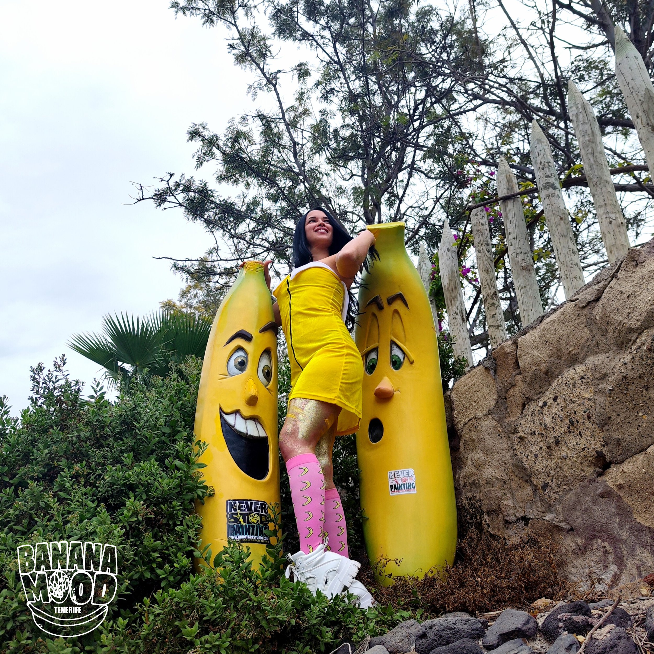 Mujer sonriente de pie entre figuras de plátano con expresiones alegres y sorprendidas en un parque, con árboles y una cerca de madera al fondo.