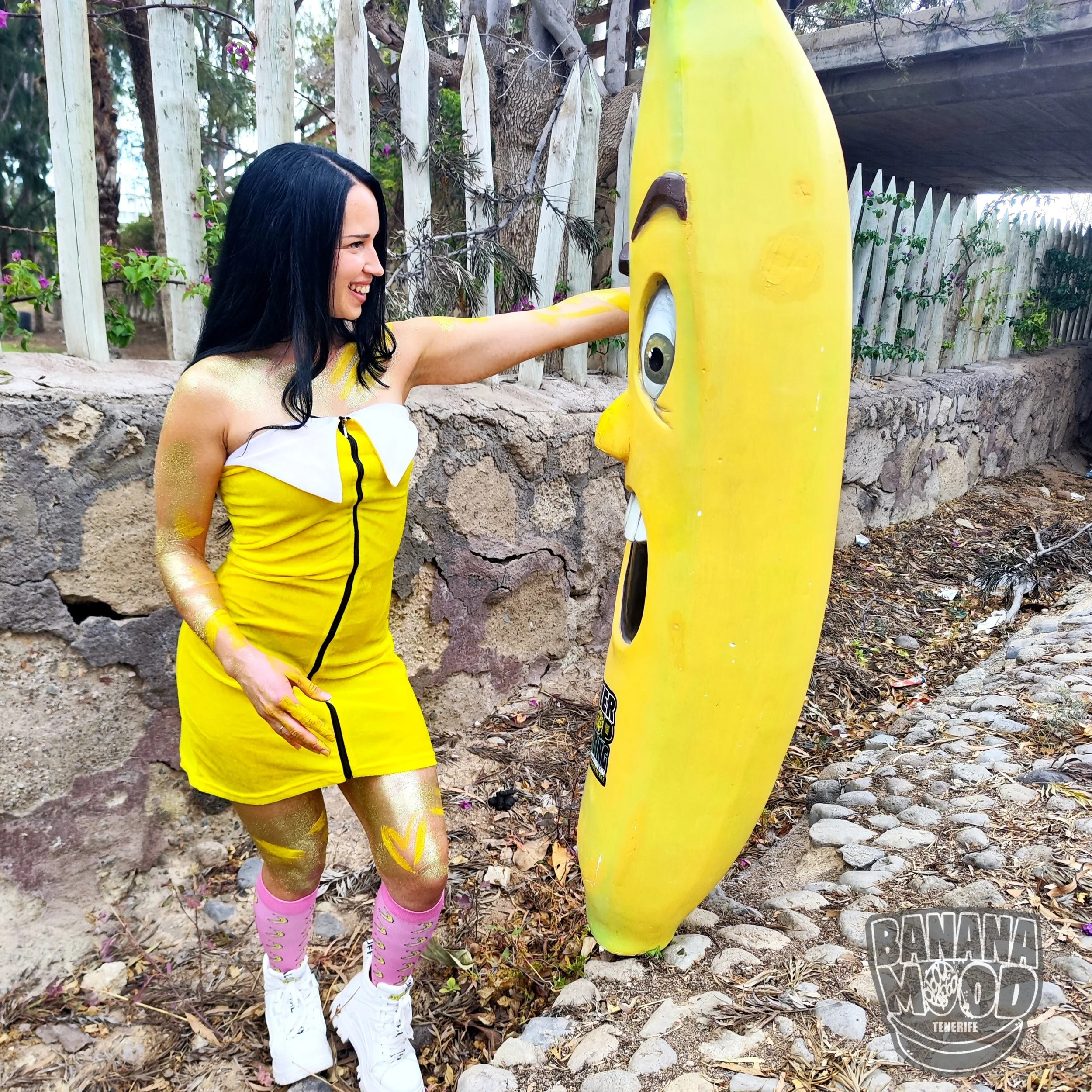 Una mujer sonriendo tocando la cara de una escultura de una banana gigante con ojos y boca, en un entorno al aire libre con cercas de madera y piedras en el suelo.