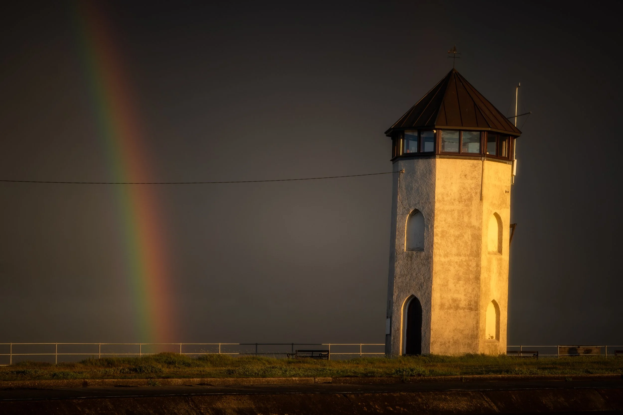 A white lighthouse with a dark metal roof stands on a grassy area at sunset, with a rainbow visible in the background against a dark sky.