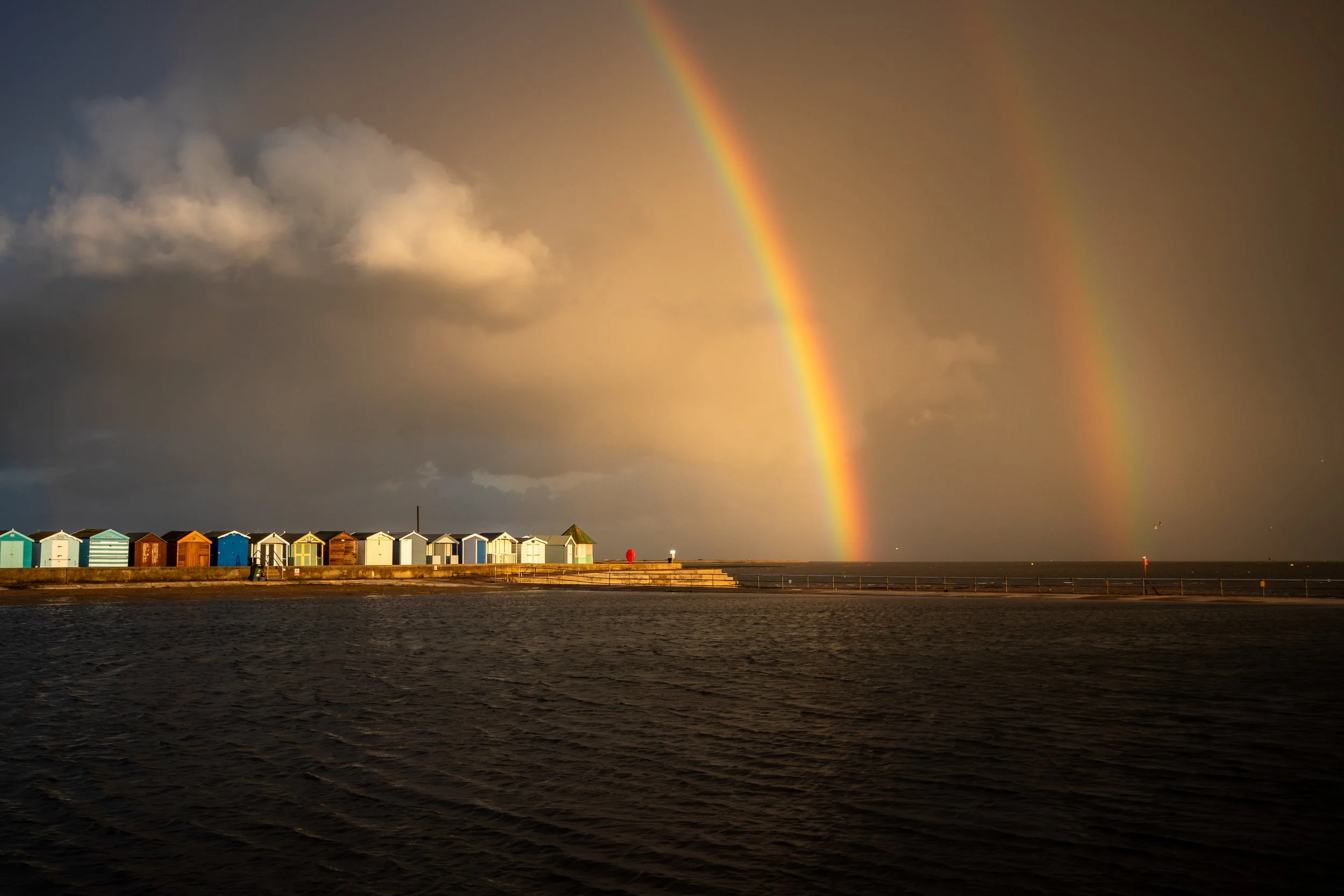 Colorful beach huts along the shoreline with a double rainbow in a cloudy sky over the water.