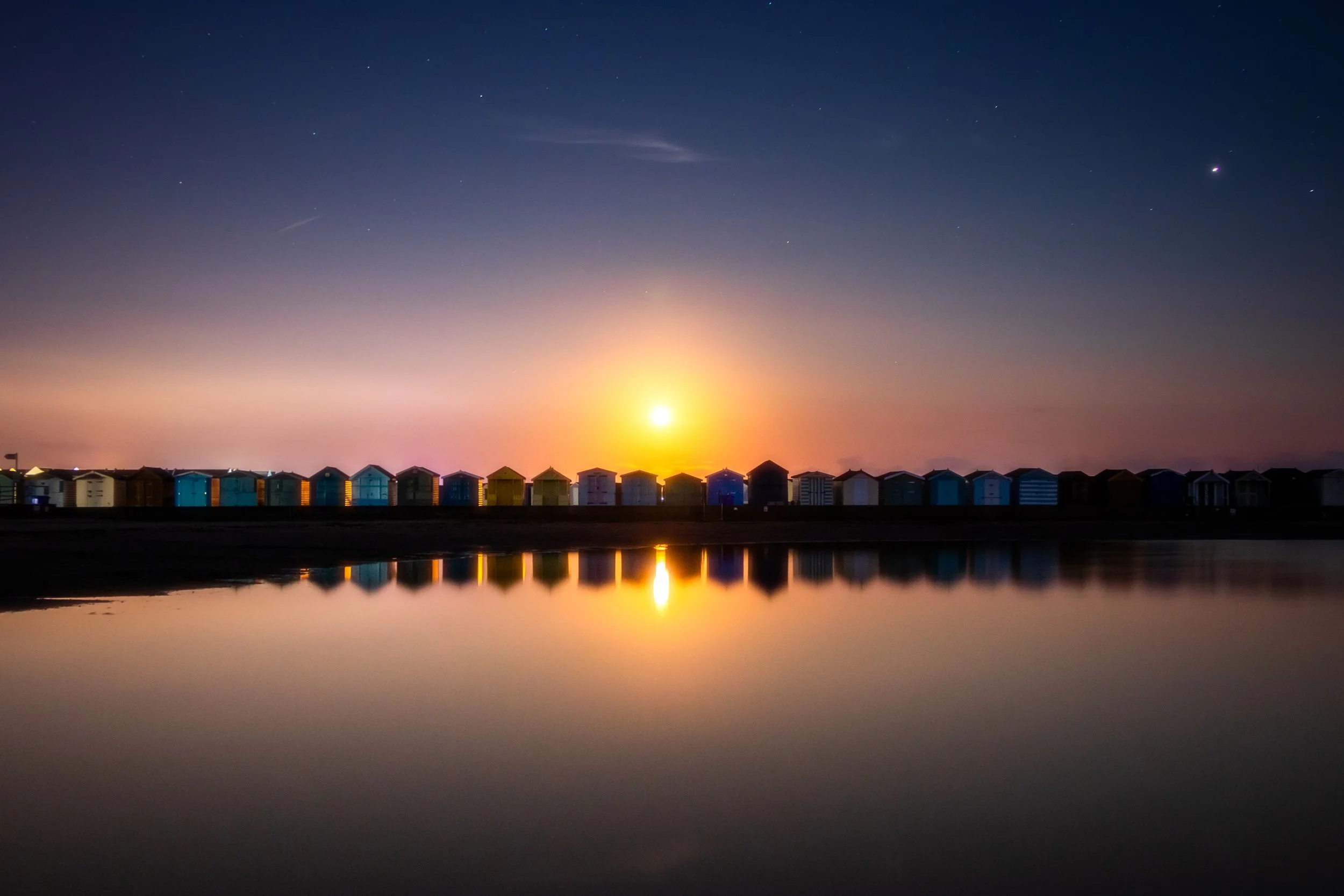 Sunset over colorful beach huts reflected in calm water under a starry sky.