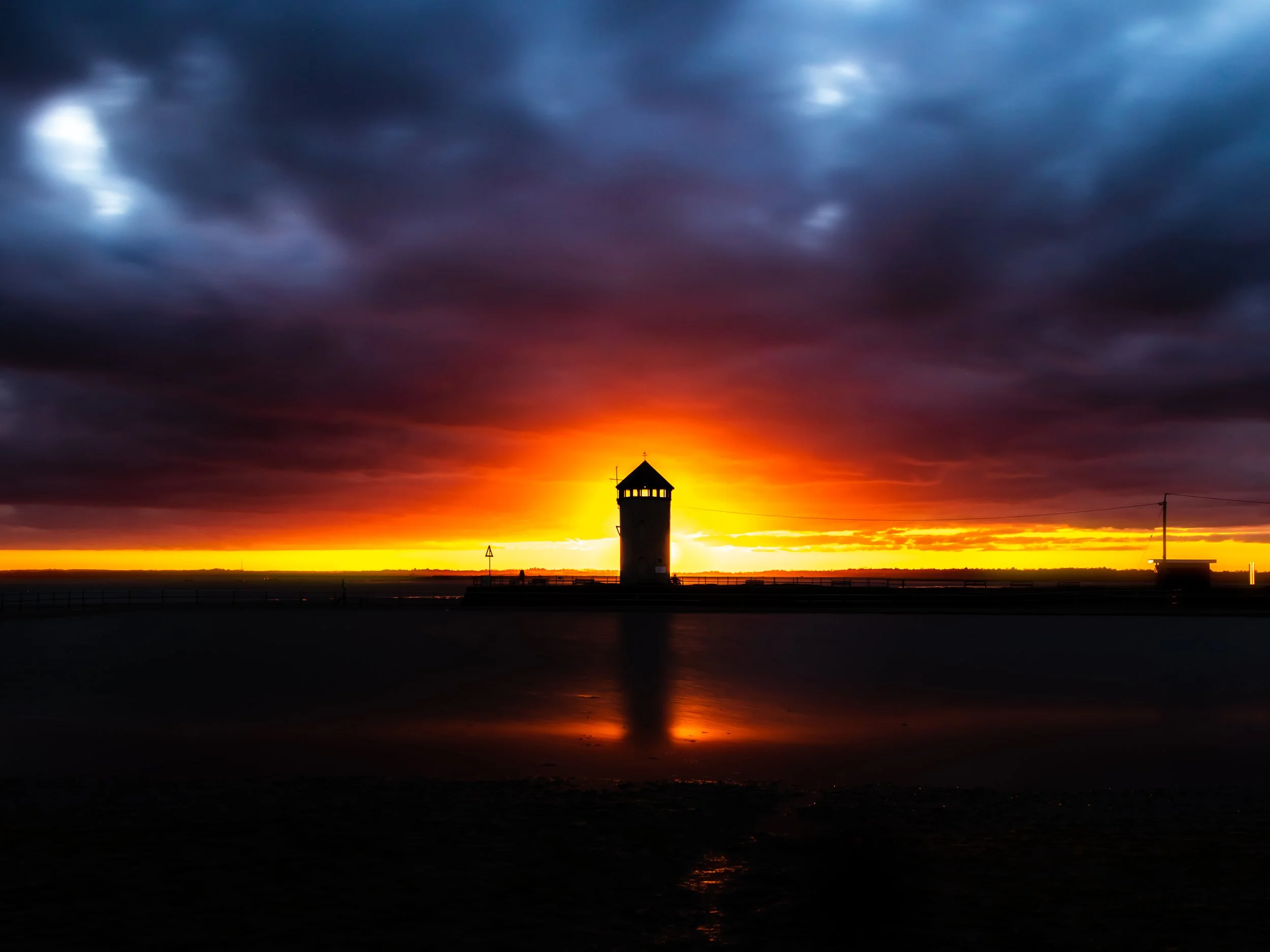 Sunset with a dark cloudy sky over a silhouetted lighthouse and its reflection on the water