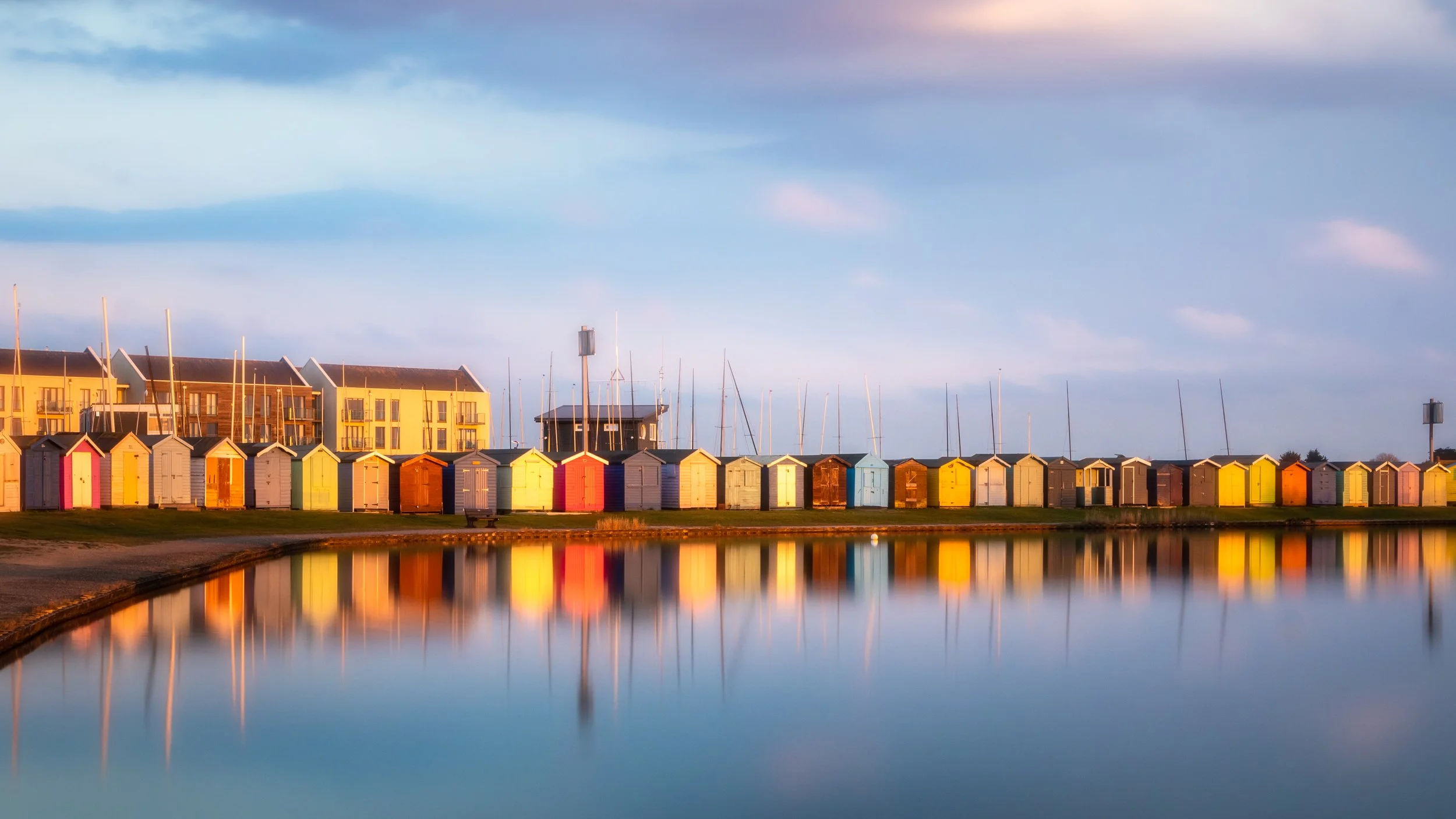 Colorful beach huts along a calm body of water with reflections, under a partly cloudy sky during sunset.