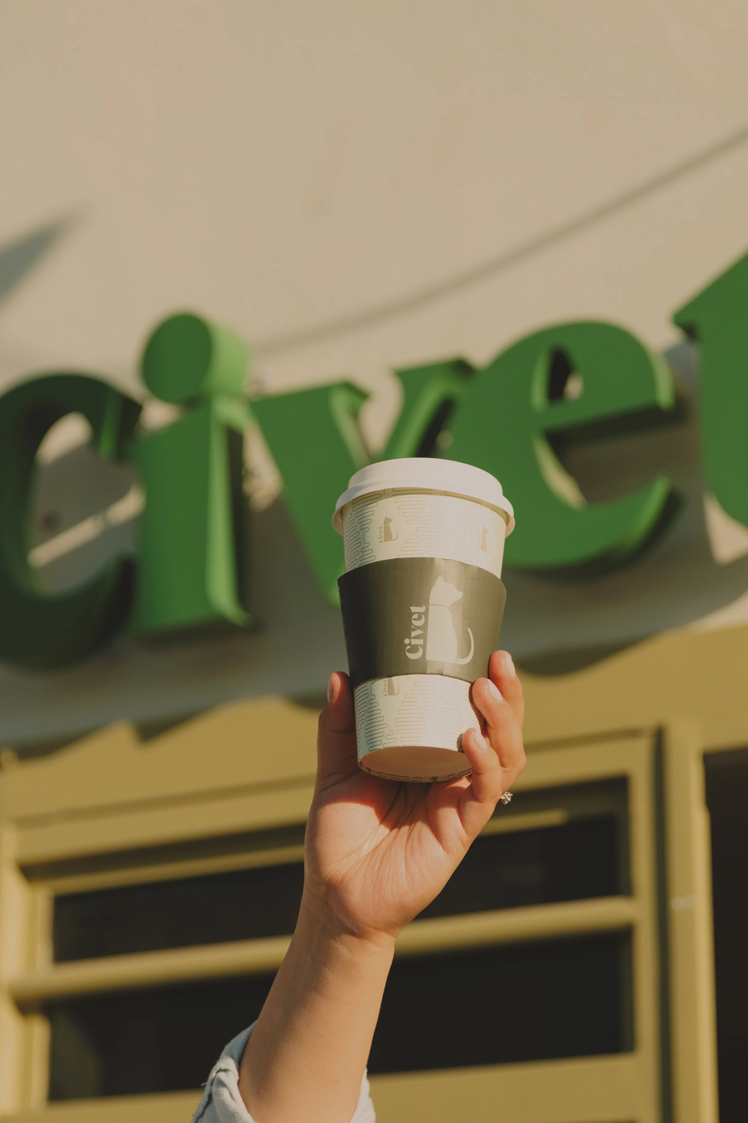 Una mano sostiene un vaso de café con tapa blanca frente a un fondo con letras verdes que forman la palabra "Civet" en un muro exterior.