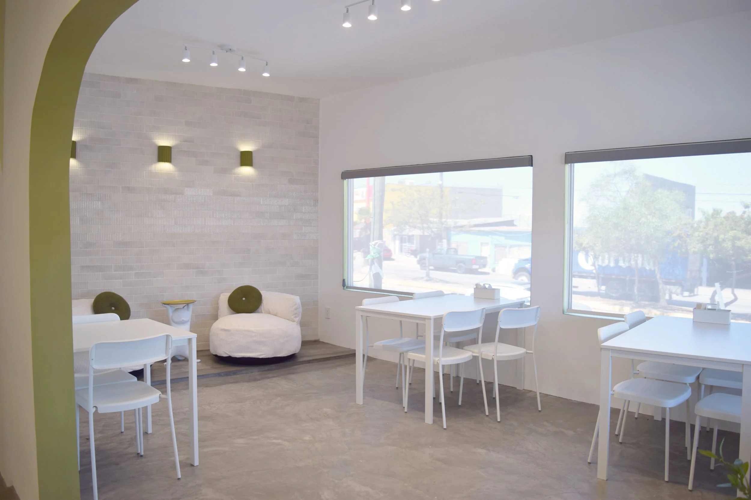 Interior de un salón con mesas blancas y sillas, paredes blancas y una pared de ladrillos claros con lámparas empotradas, ventanas grandes que dejan entrar luz natural, y una zona de descanso con sillones blancos y cojines verdes.