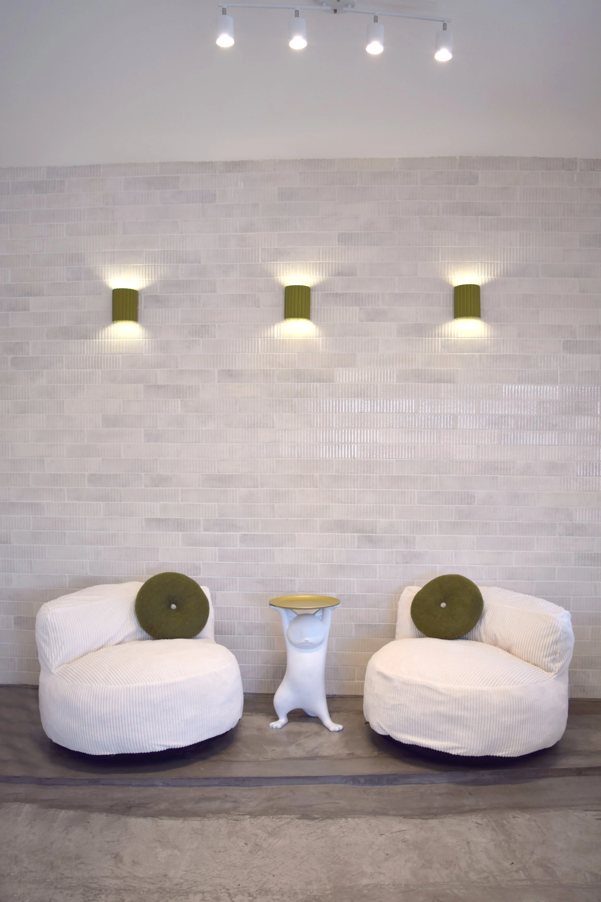 Sala de descanso con dos sillones blancos de felpa, cojines verdes, un lado de una mesa decorativa en forma de animal y una pared con luces y accesorios de pared en color verde.