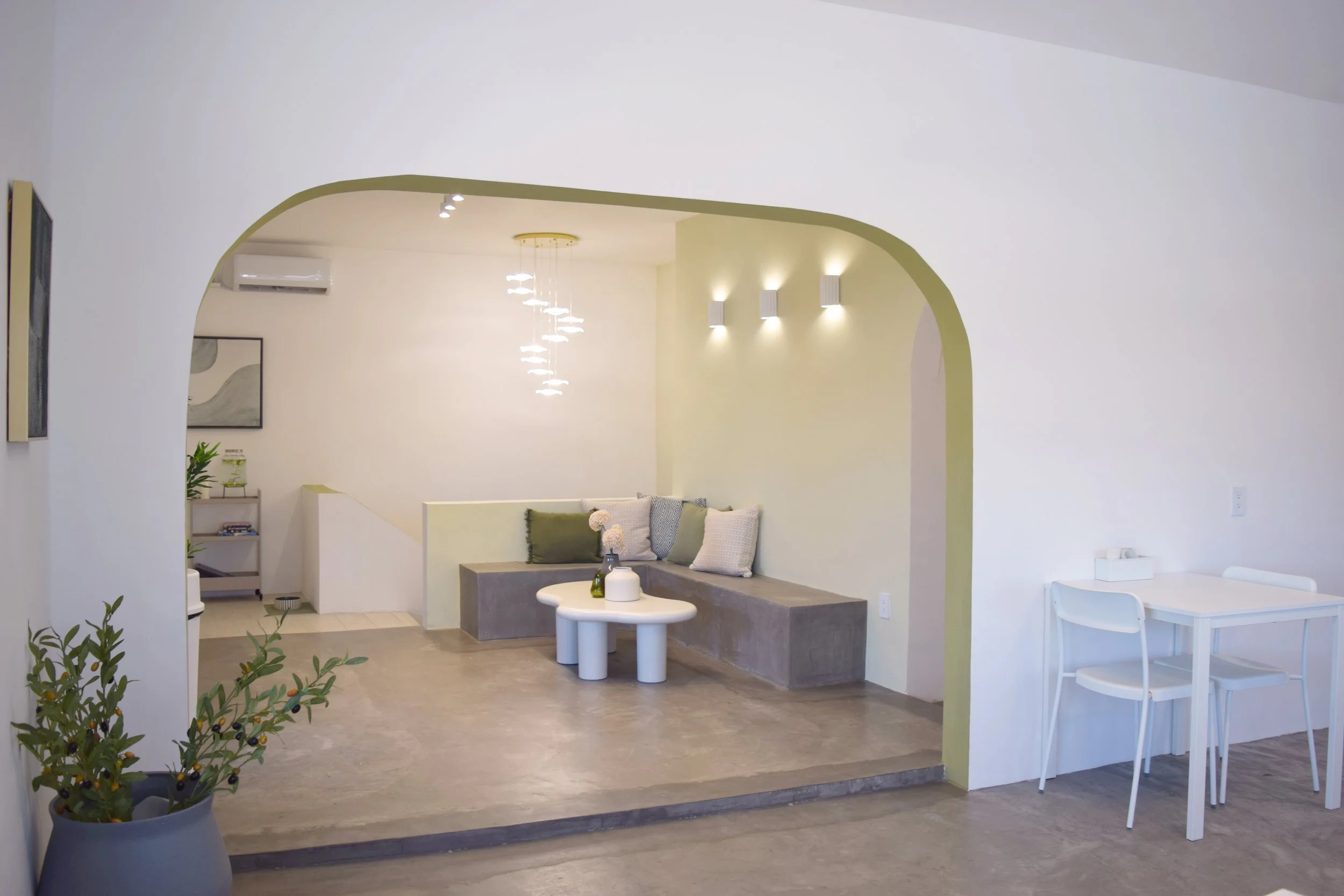 Sala moderna con un banco de concreto, cojines blancos y verdes, lámpara moderna, paredes blancas y una pequeña mesa blanca con decoraciones.