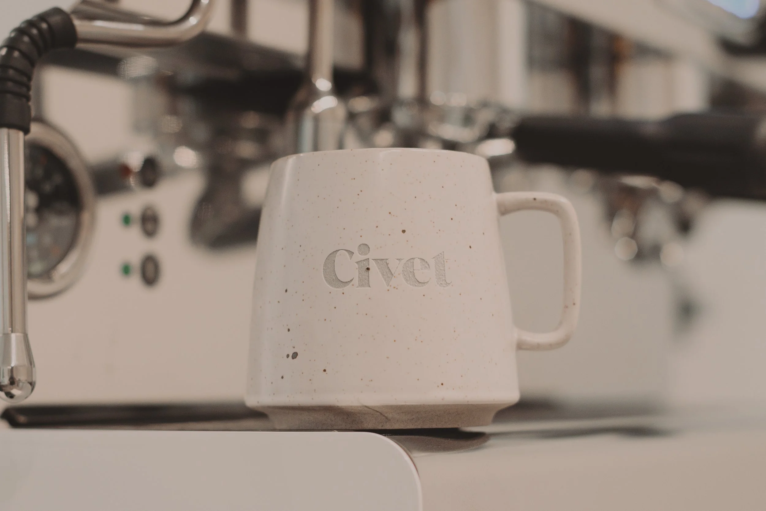 Taza de cerámica blanca con marca 'Civel' en un fondo de maquinaria de barista.
