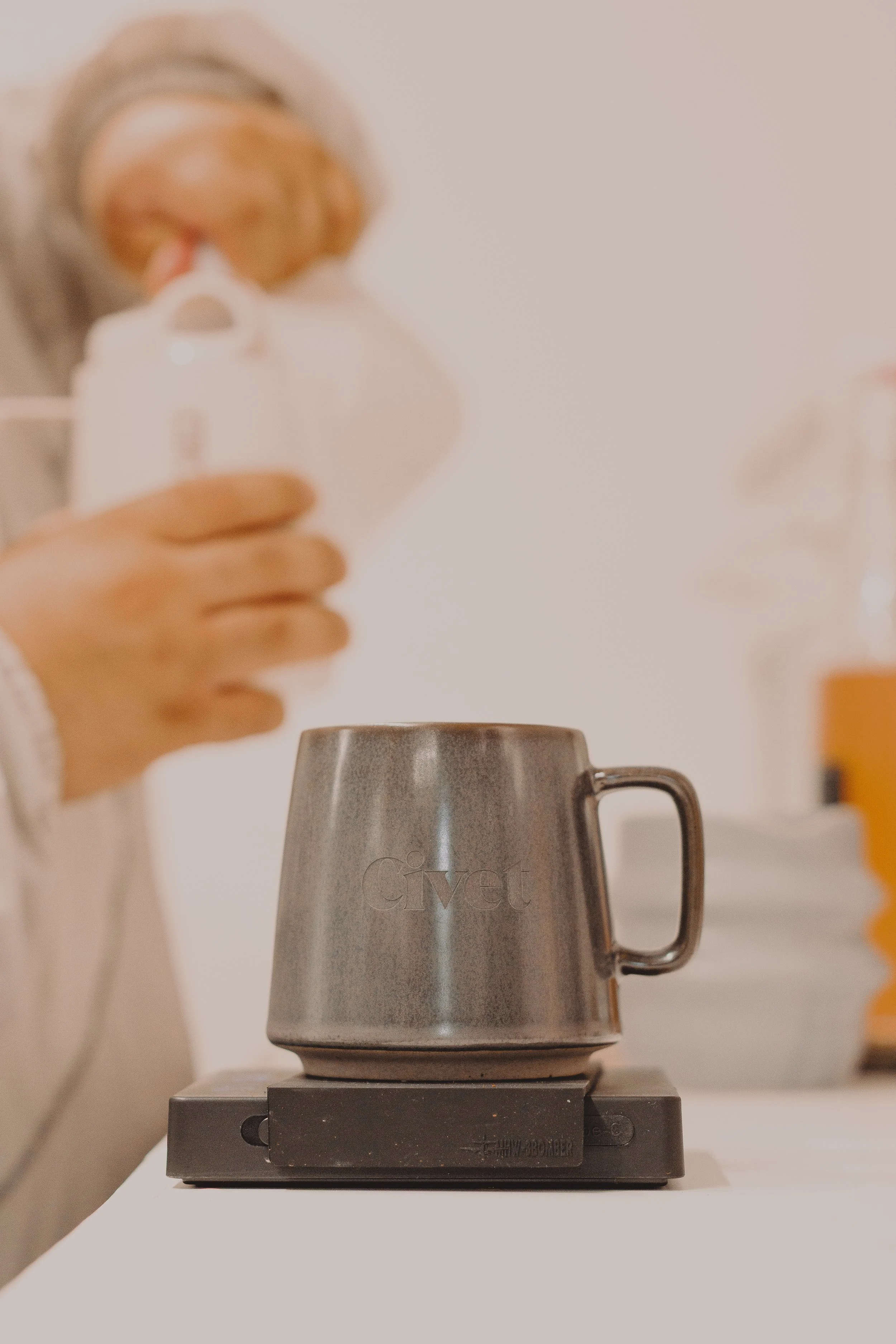 Alguien agrega leche a un vaso de metal en una balanza, con una taza y otros objetos en el fondo.