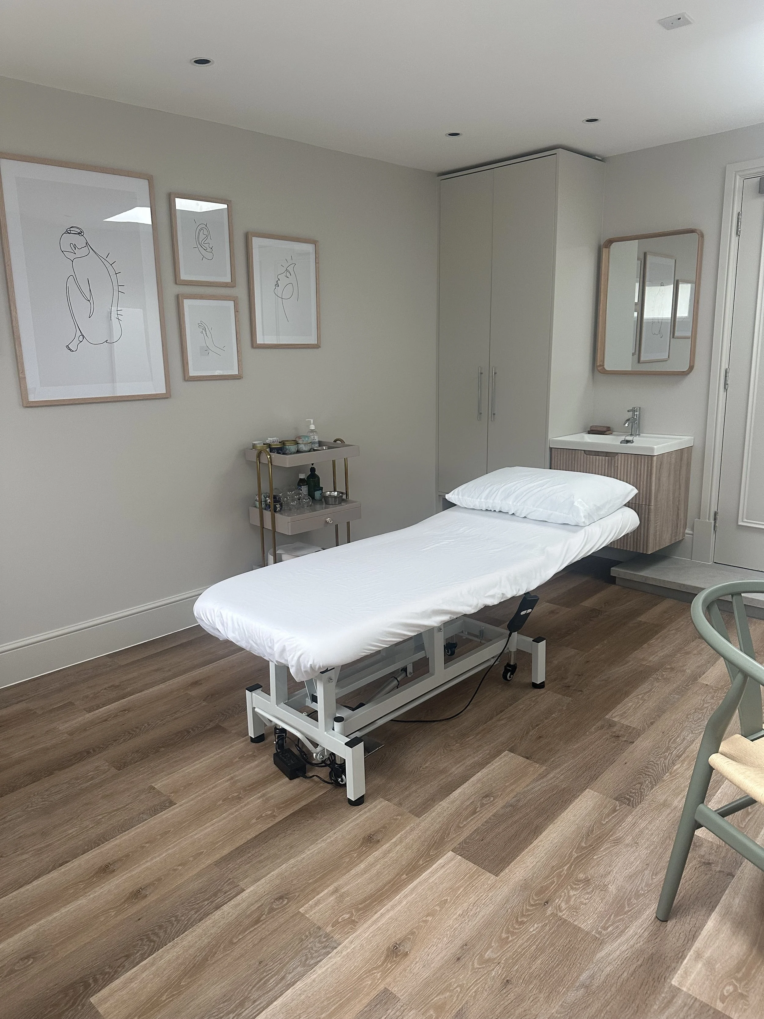 A medical treatment room with a hospital bed, small sink with a mirror, framed line art drawings on the wall, and a rolling cart with supplies.