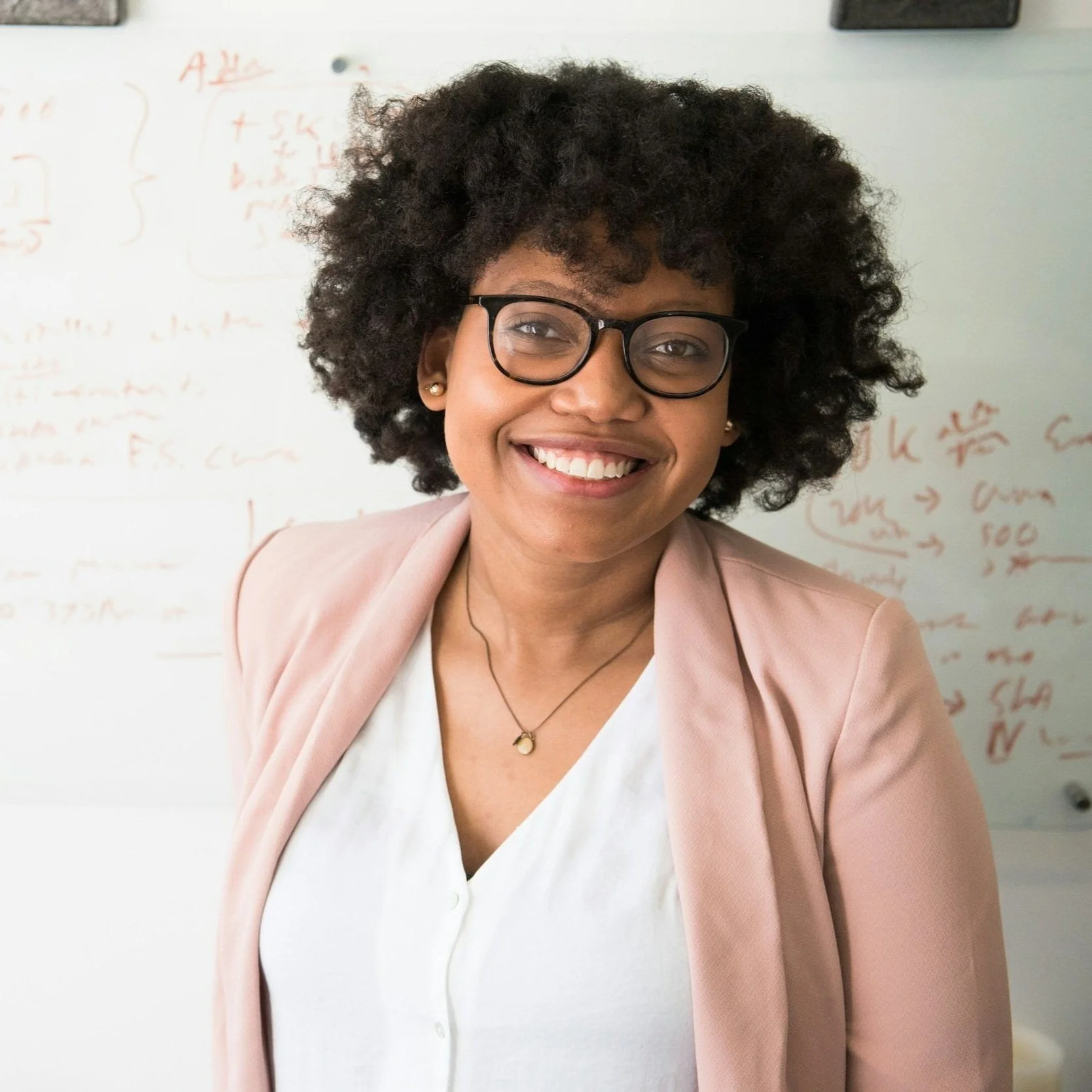Smiling woman with curly hair, glasses, and a pink blazer standing in front of a whiteboard with red writing.