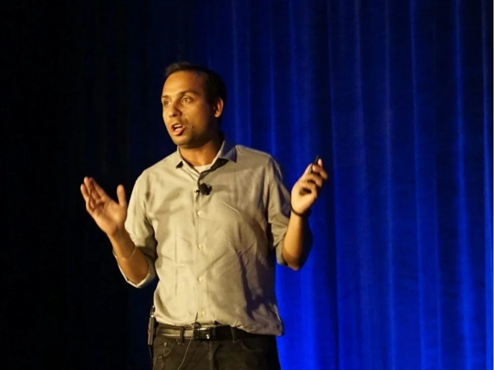 Man giving a presentation on stage with blue curtains in the background.