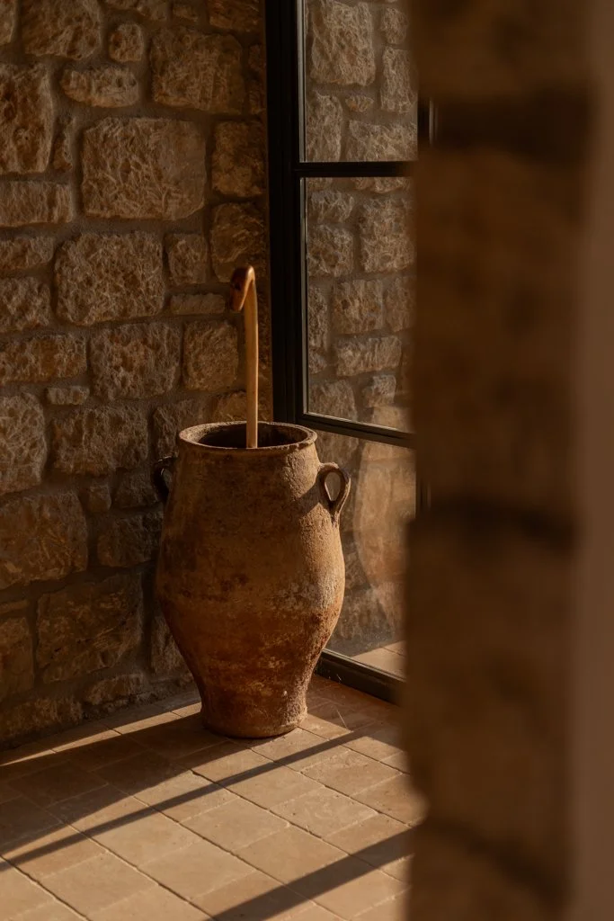 Ancient terracotta water jar with a wooden dipper inside, positioned next to a window with stone wall background, casting shadow on tiled floor.