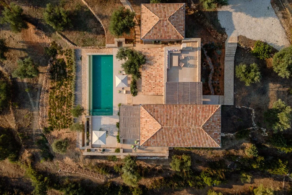 Aerial view of a house with a swimming pool, surrounded by trees and landscape, with tiled roofs and outdoor seating areas.