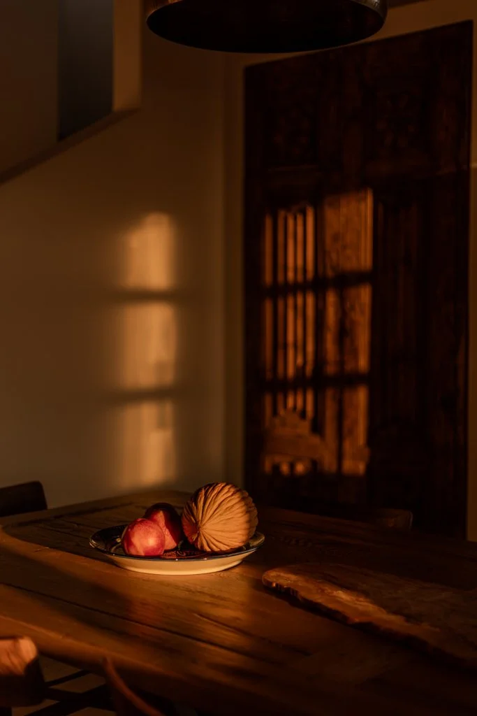 A dimly lit dining table with a plate holding a pumpkin and an apple, a wooden panel or cutting board, and shadows cast by sunlight through a window.