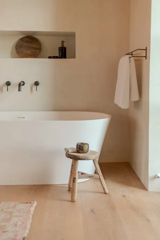 A minimalist bathroom featuring a white bathtub, a small wooden stool with a candle on top, towel hanging on a wall-mounted rack, and a decorative shelf with a round wooden object and a black bottle.