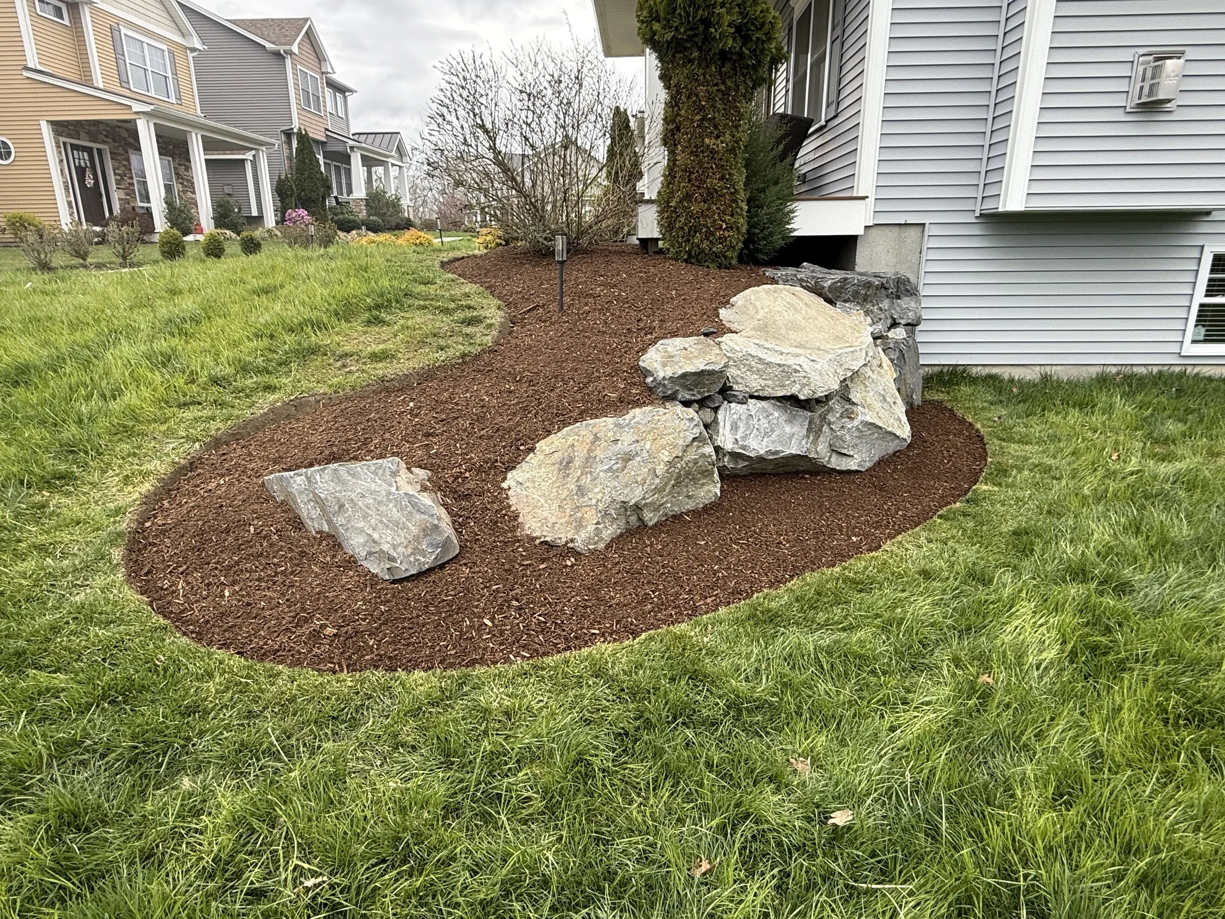 A landscaped yard with green grass, mulch, large rocks, and trimmed bushes in front of townhouses with vinyl siding.