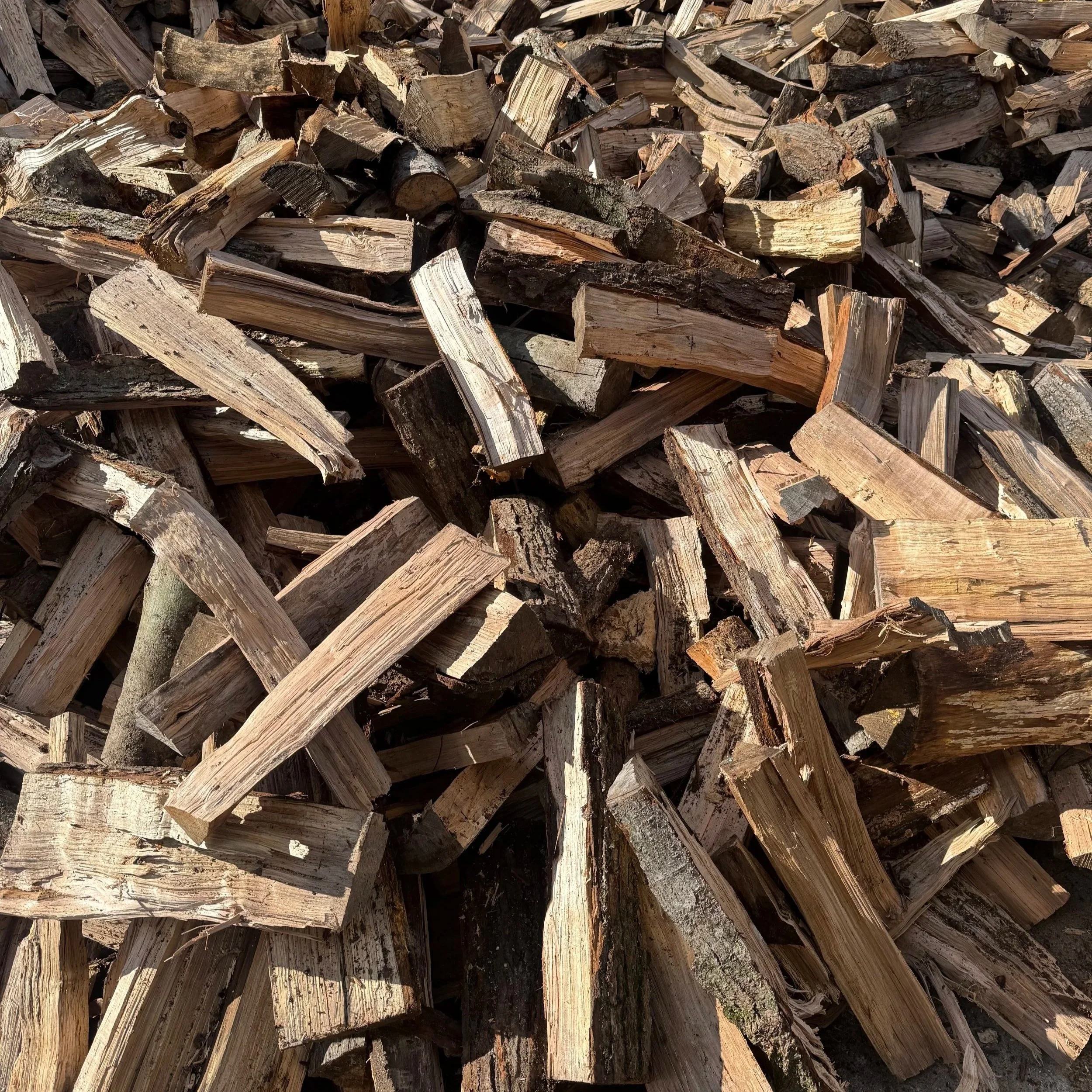 Stacked split firewood logs with rough, weathered surfaces, some showing bark and wood grain, piled outdoors in sunlight.