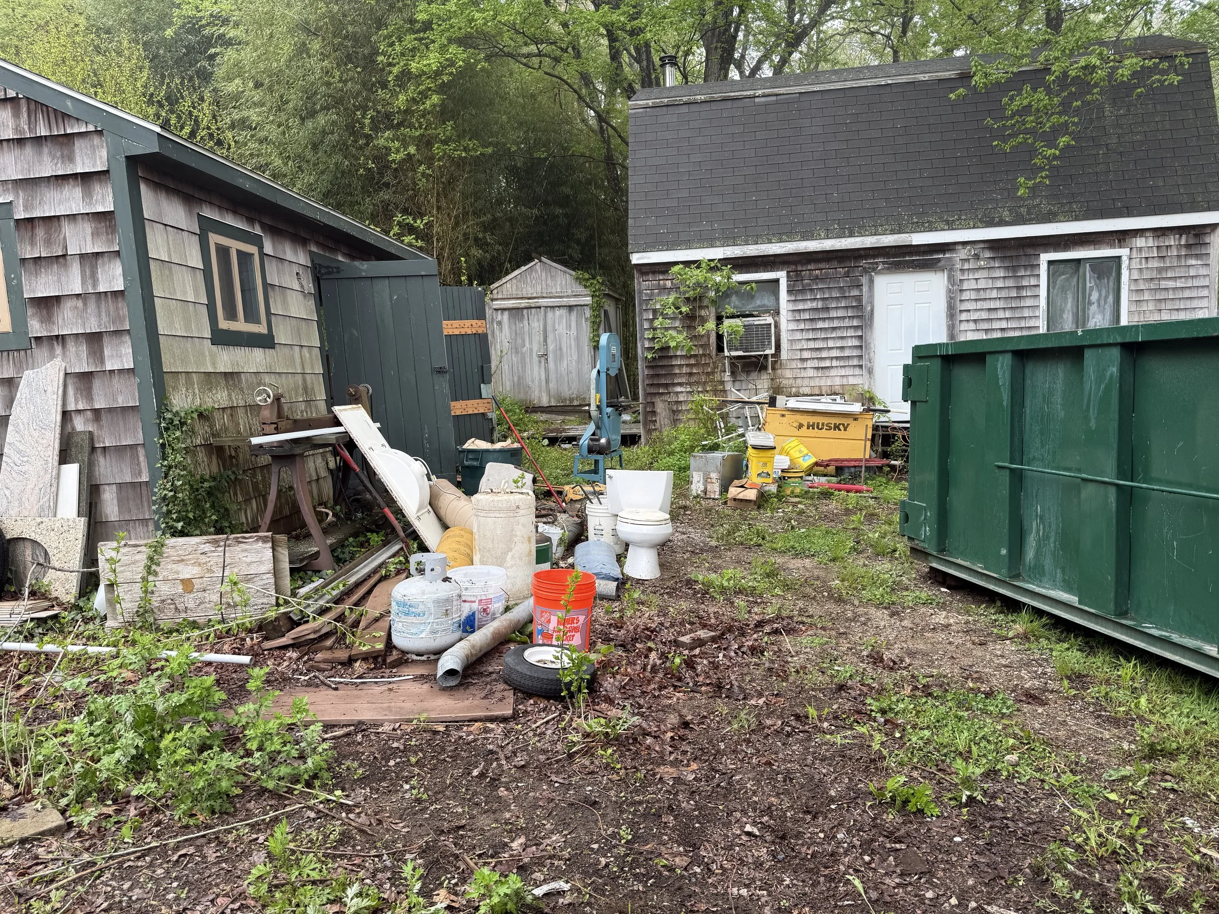 Backyard cluttered with various items including buckets, pipes, tires, and construction materials, with two small houses and greenery in the background.