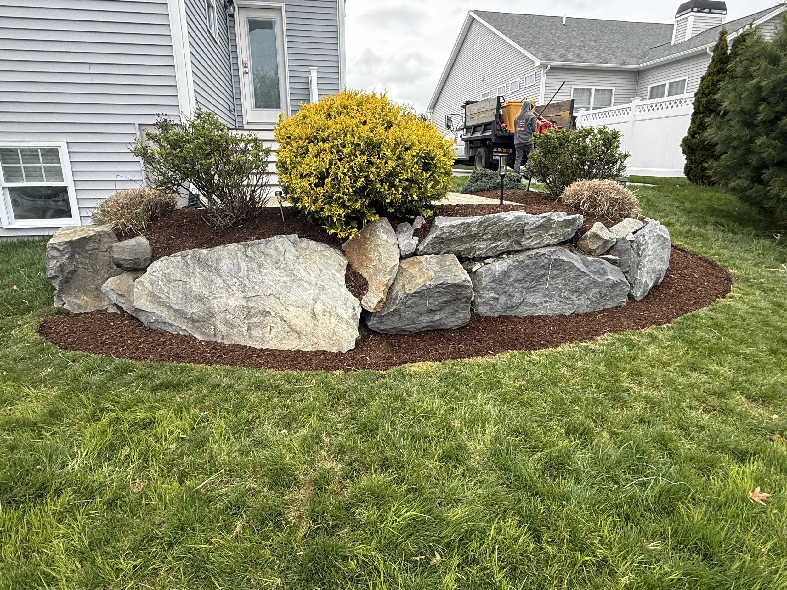 Residential backyard with a rock garden, bushes, and a person in a hoodie working on a truck.