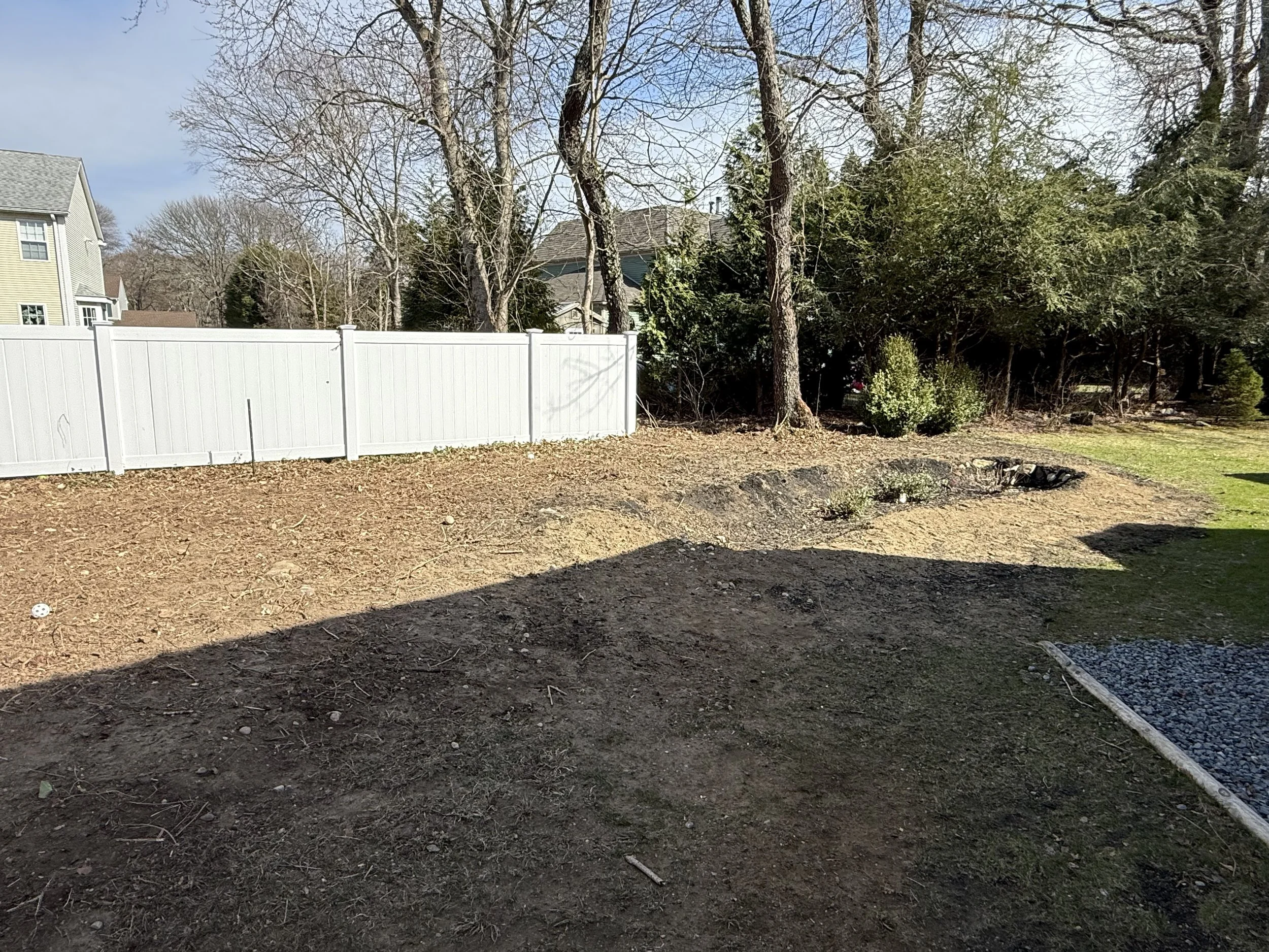 A backyard with a white privacy fence, some large trees with bare branches, some greenery, and a house in the background. The foreground shows bare soil with some small plants and garden edging.