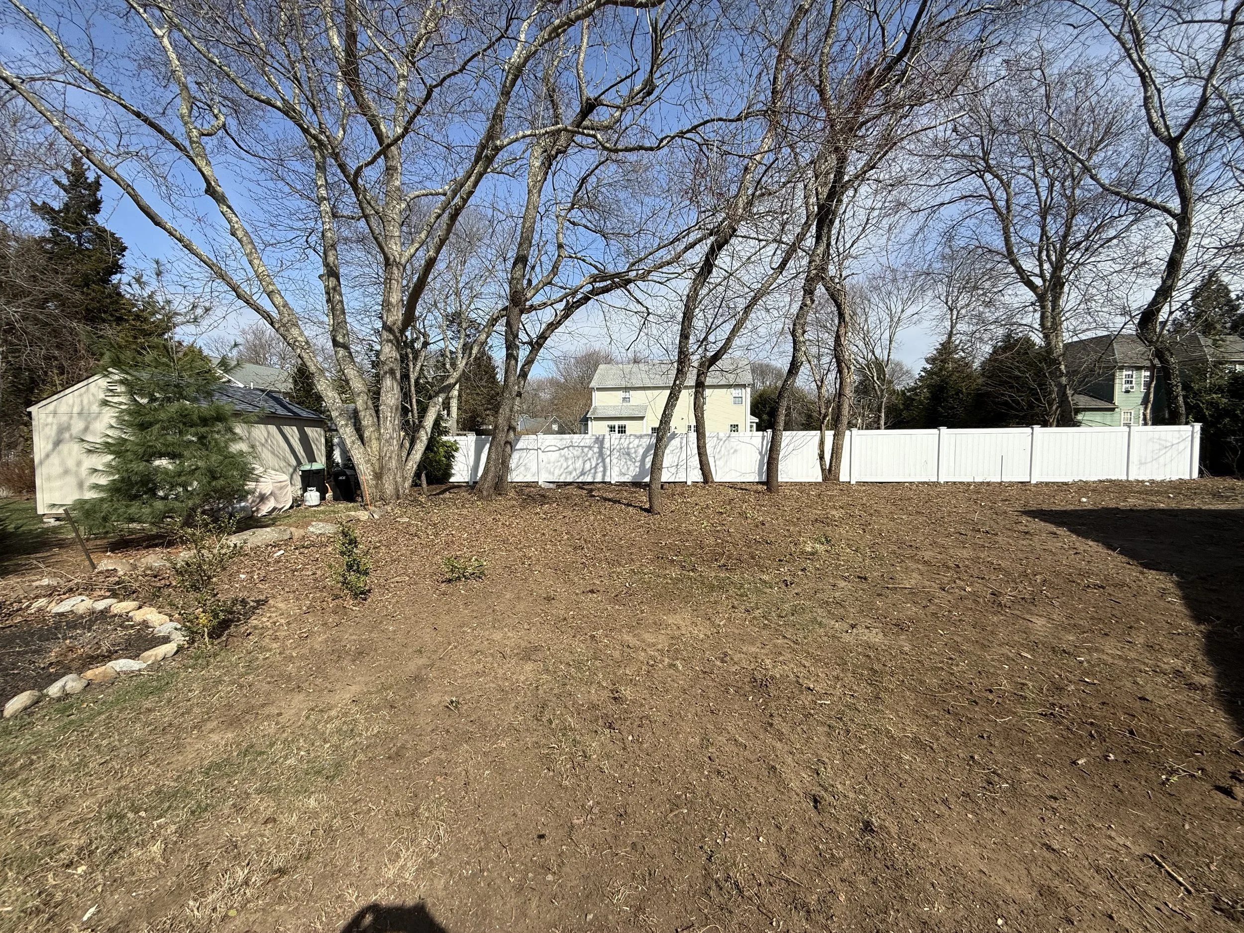 Backyard with bare trees, a white fence, a small shed, and a beige house in the background under a blue sky.