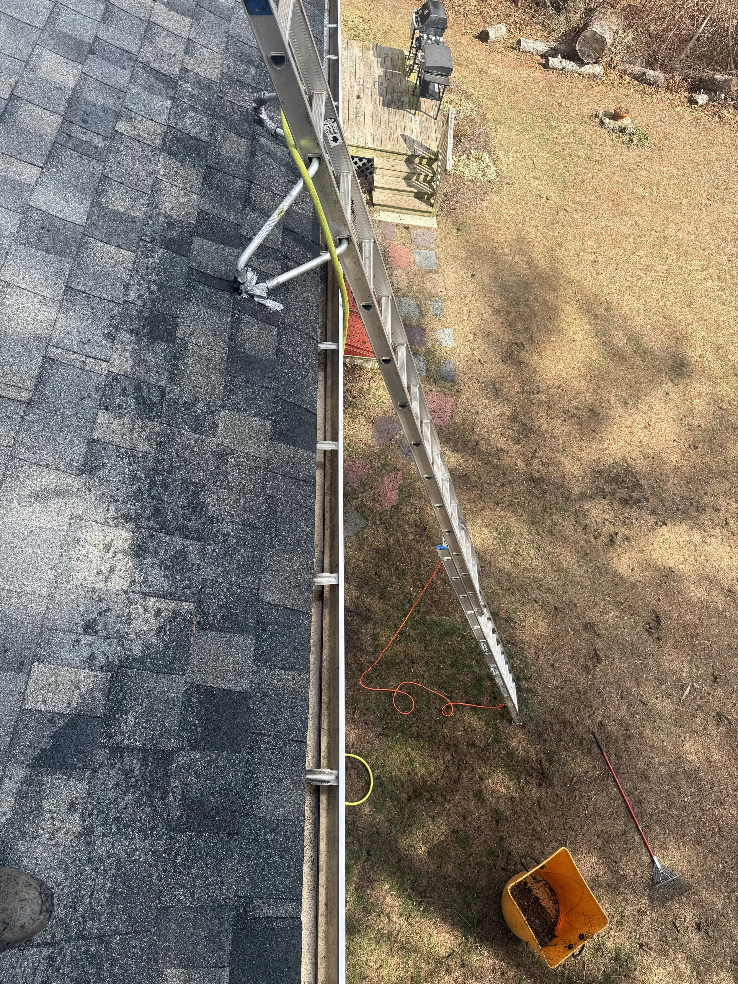 A close-up view of a roof with black asphalt shingles being installed. A metal ladder extends from the ground to the roof, with an orange extension cord running next to it. On the ground, there is a yellow bucket filled with some material, possibly r