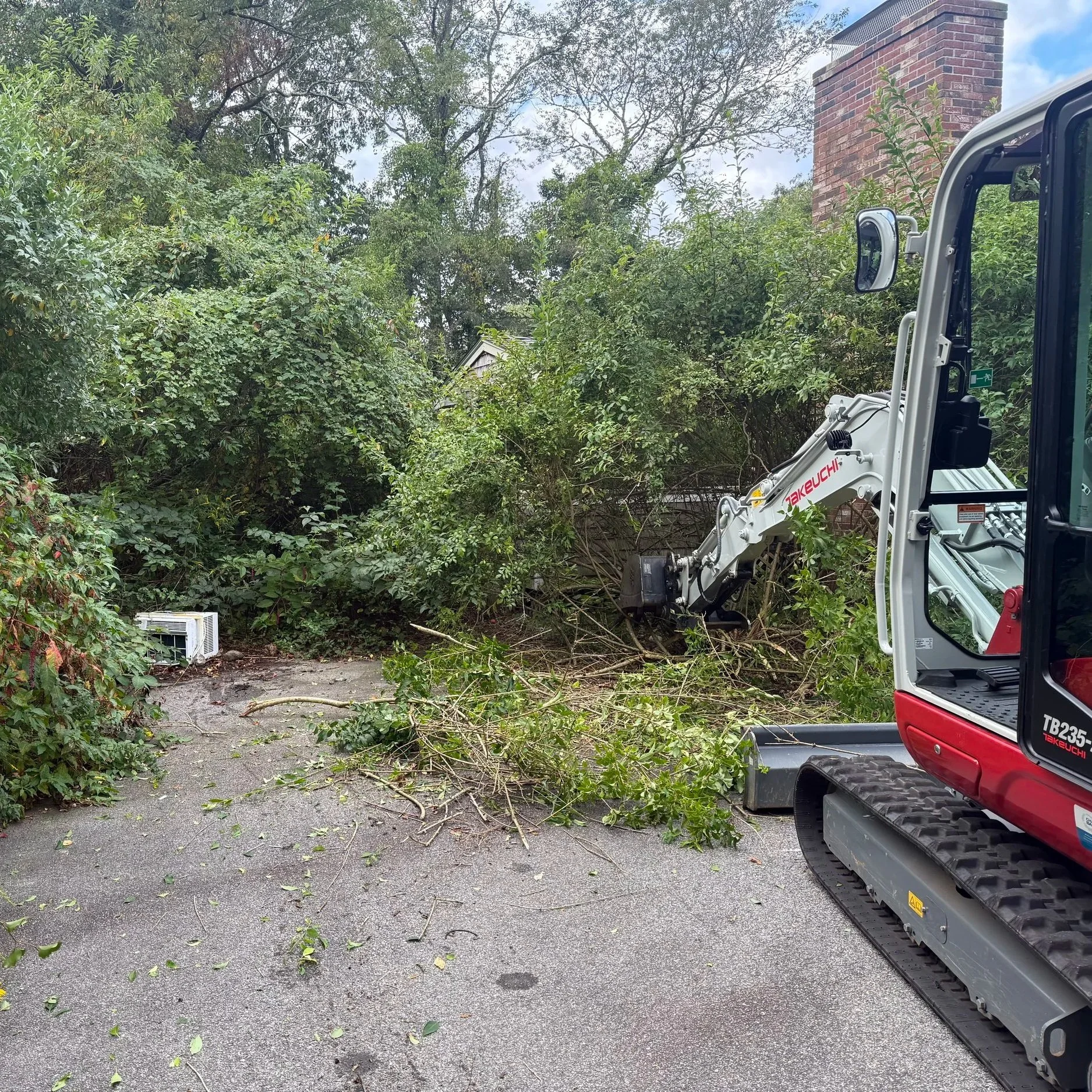 A small excavator clearing fallen branches and debris from a driveway surrounded by dense green trees. A brick house chimney is visible in the background.