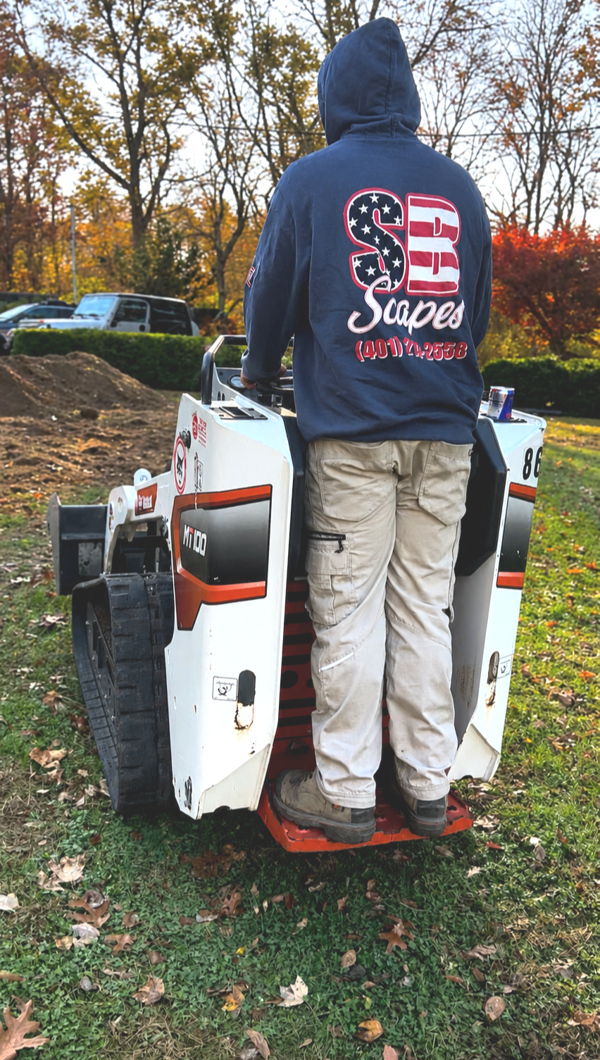 A person with a hoodie standing on an industrial machine outdoors during autumn, with trees with fall-colored leaves in the background.