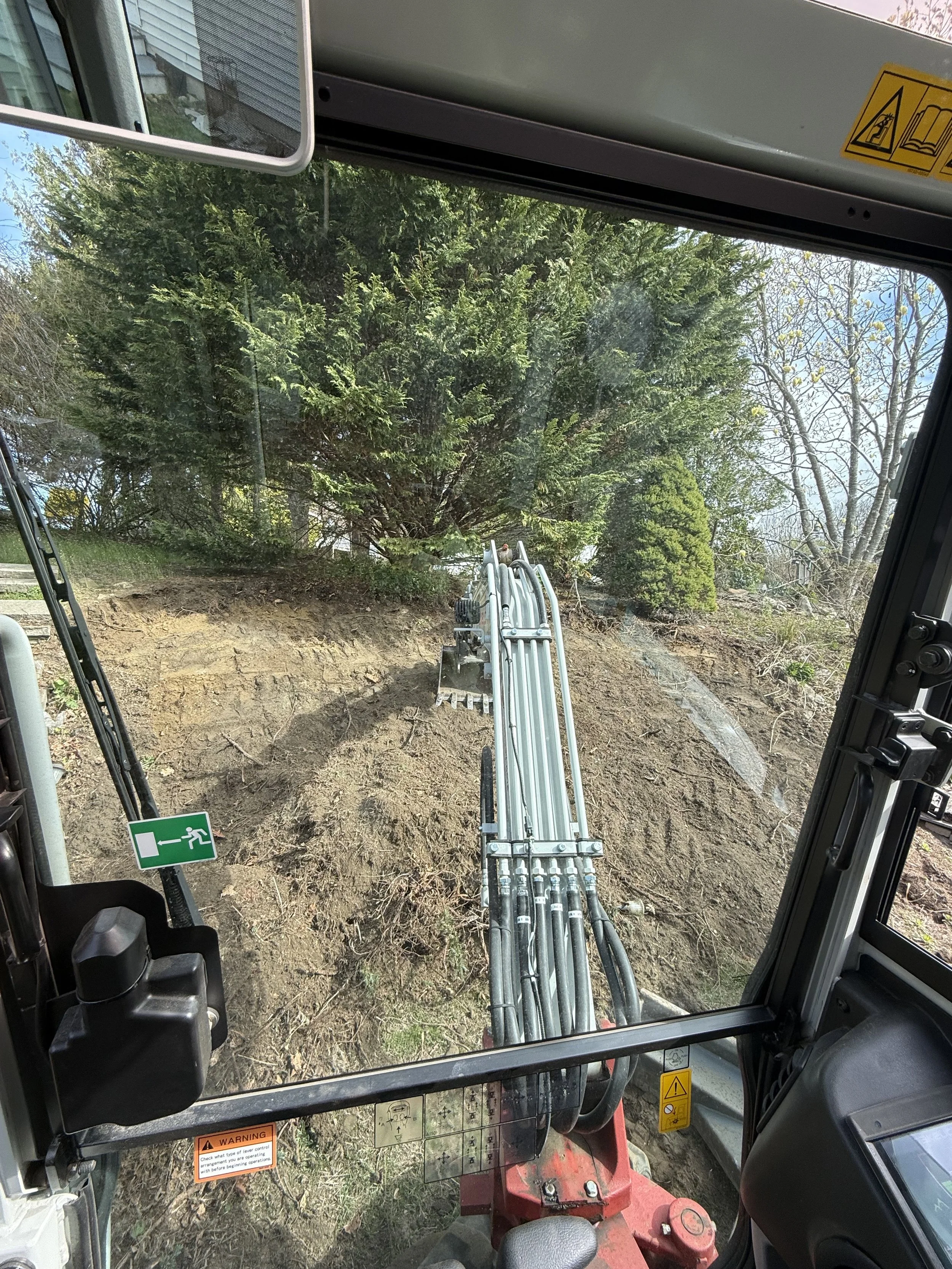 Inside the cabin of a construction vehicle, looking out onto a dirt work site with trees and foliage in the background.