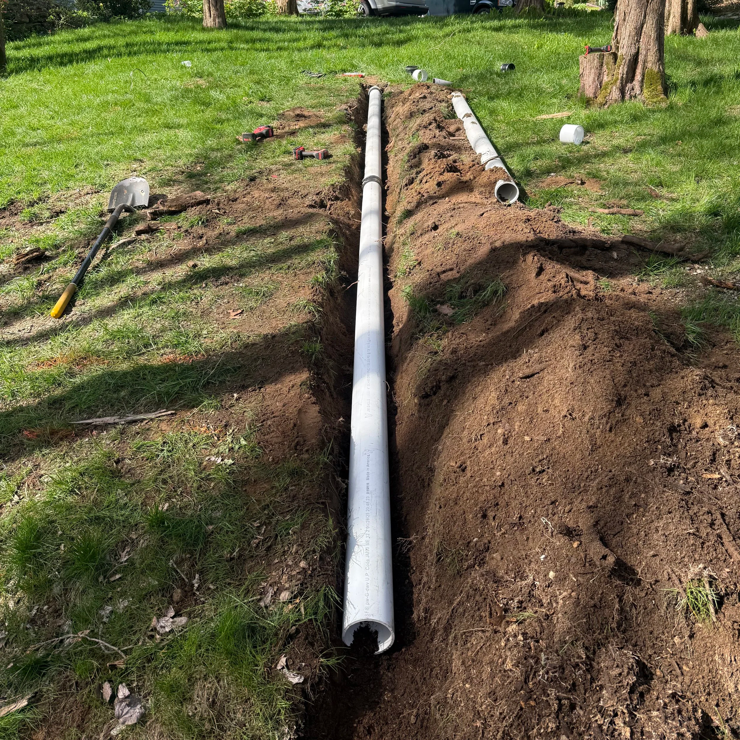 A trench dug in a grassy backyard with white pipes laid inside. Gardening tools and debris are scattered around the trench, and trees are visible in the background.