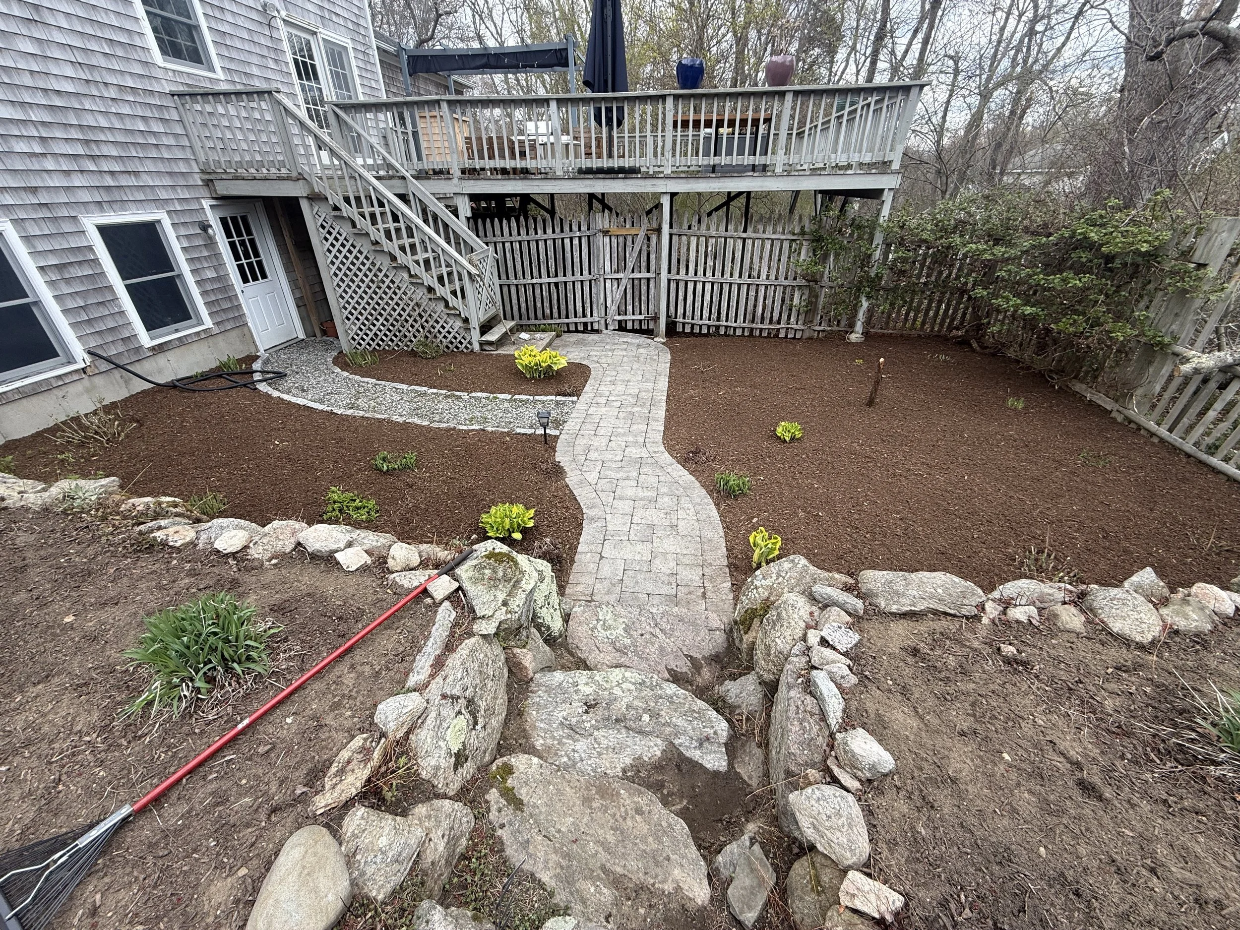 Backyard with a wooden deck, potted plants, a stone pathway, newly planted flowers, and a garden rake on the ground.
