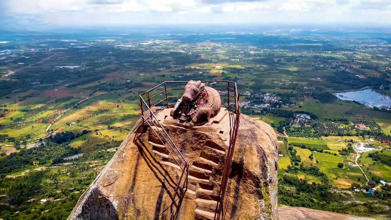 A stone sculpture of an elephant at the top of a tall rock formation, with stairs and railing leading up to it, set against a vast green landscape and cloudy sky.