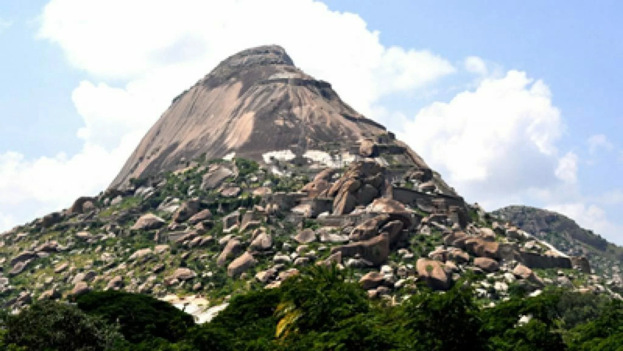 A mountain with a rocky, sloped face covered in large rocks and some green vegetation at its base, under a partly cloudy sky.