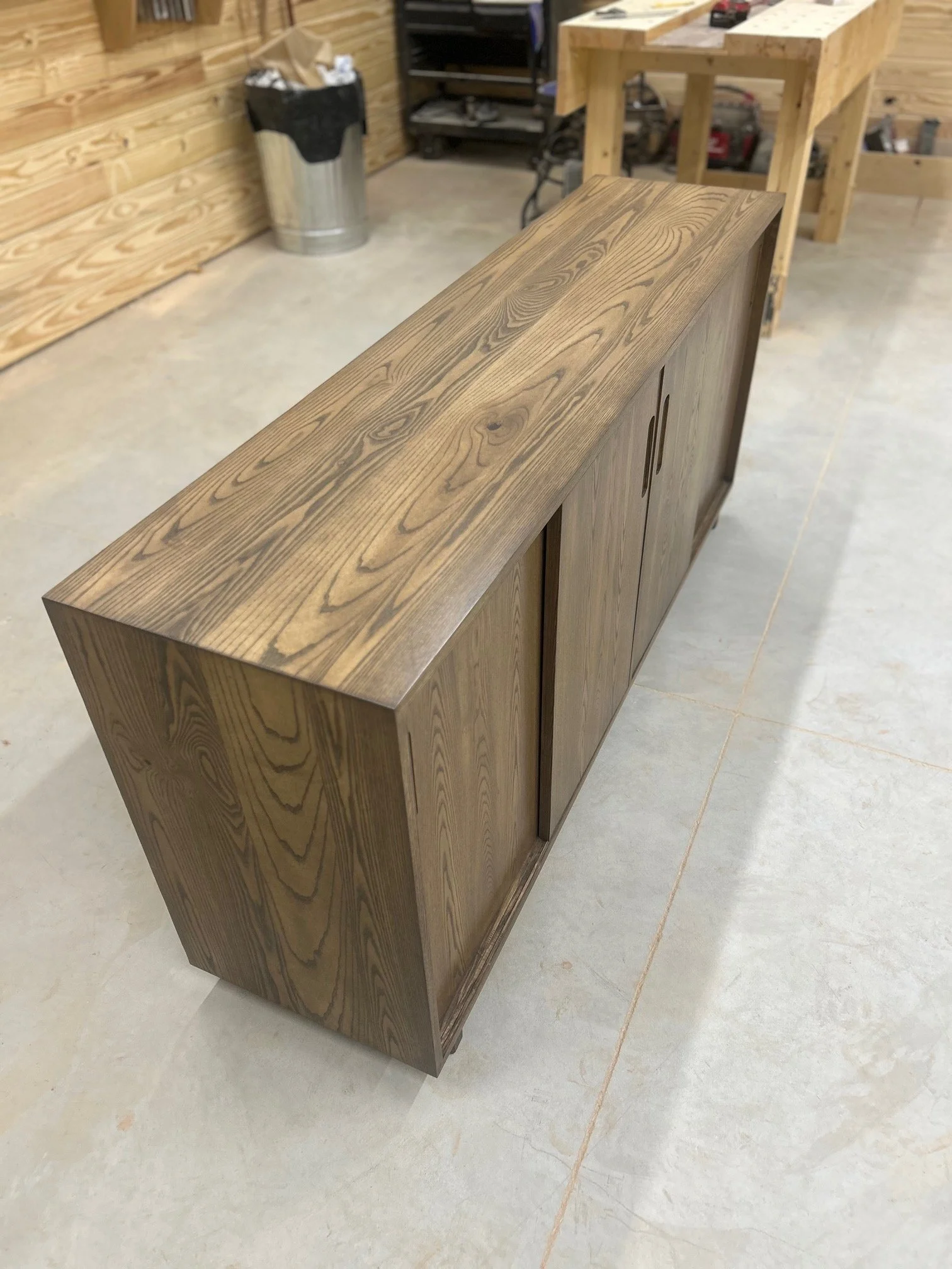 A wooden sideboard with a natural finish, placed on a concrete floor in a workshop or woodworking shop.