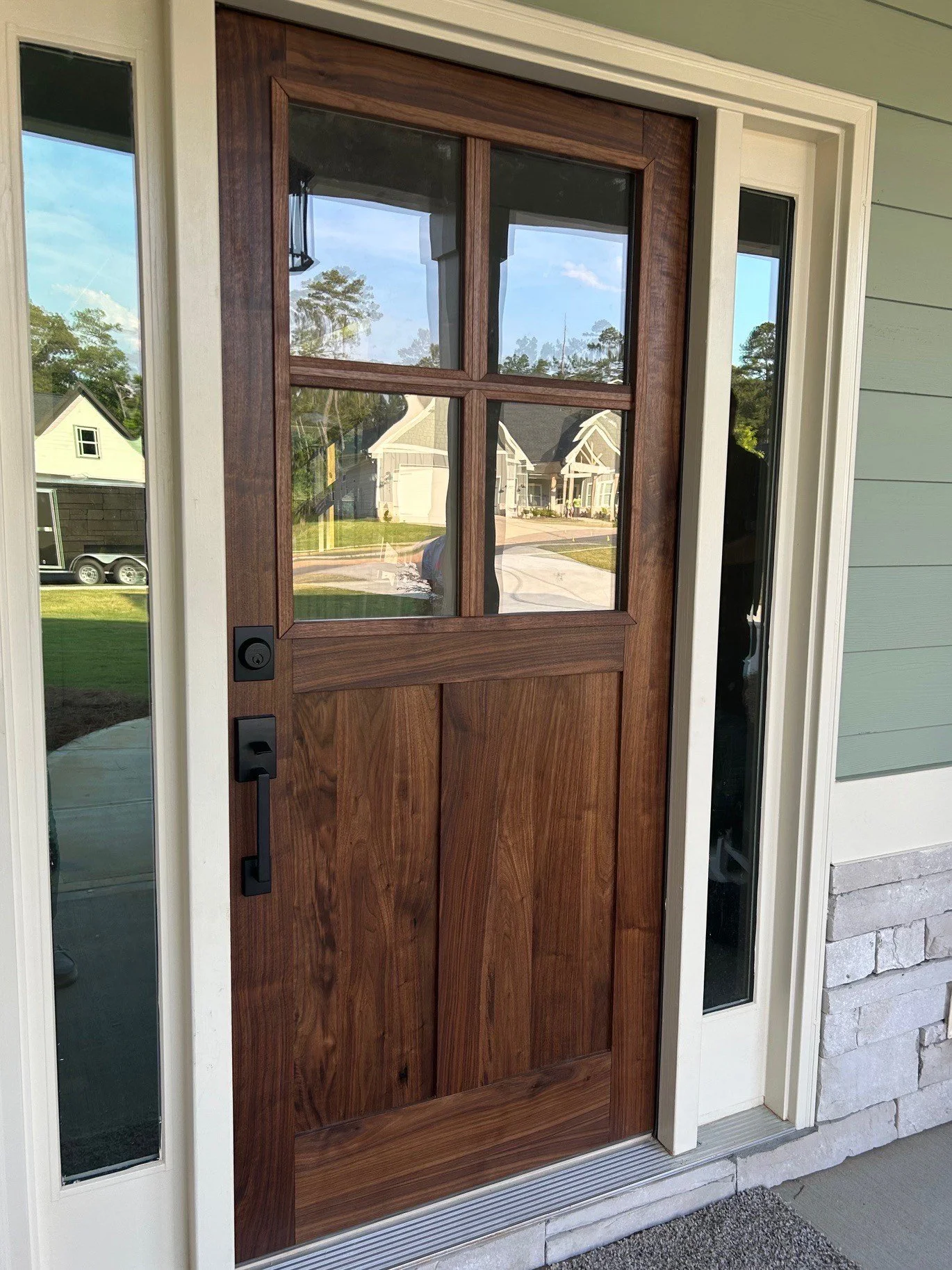 Front door with a wooden finish and four glass panels, flanked by sidelight windows, on a house with light green siding and stone foundation.