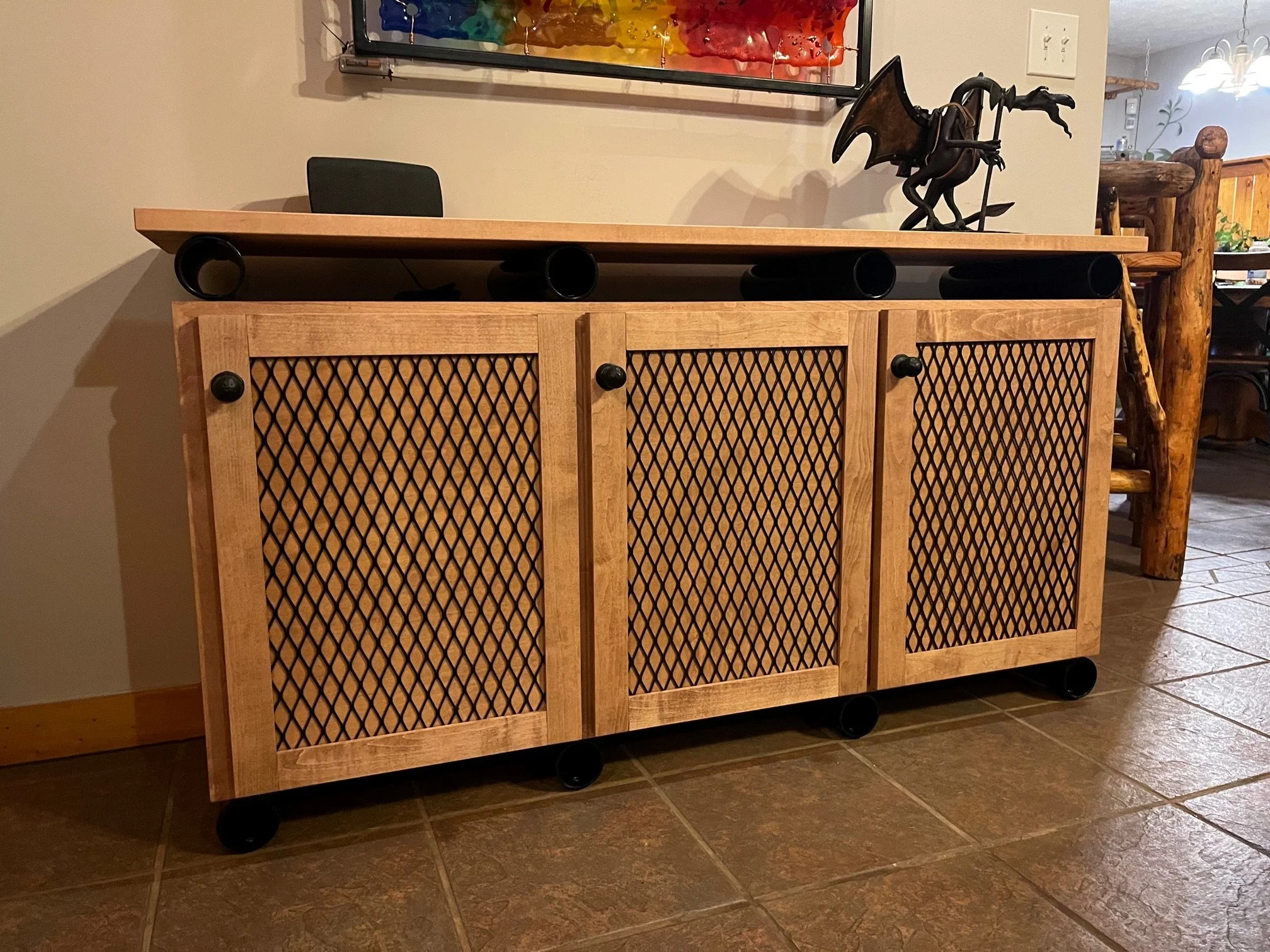 Wood and metal cabinet with lattice doors, featuring three black pipes and a metal dragon sculpture on top, in a room with tiled floor and other furniture.