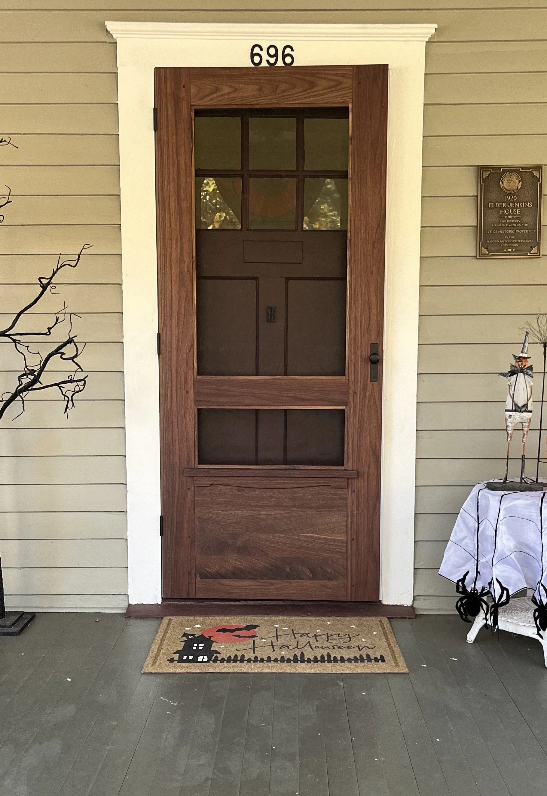 Front door of a house with a doormat that says "Happy Halloween" and has a black haunted house, a pumpkin, and a black cat. The house number 696 is above the door. There is a plaque on the wall to the right of the door and Halloween decorations, incl
