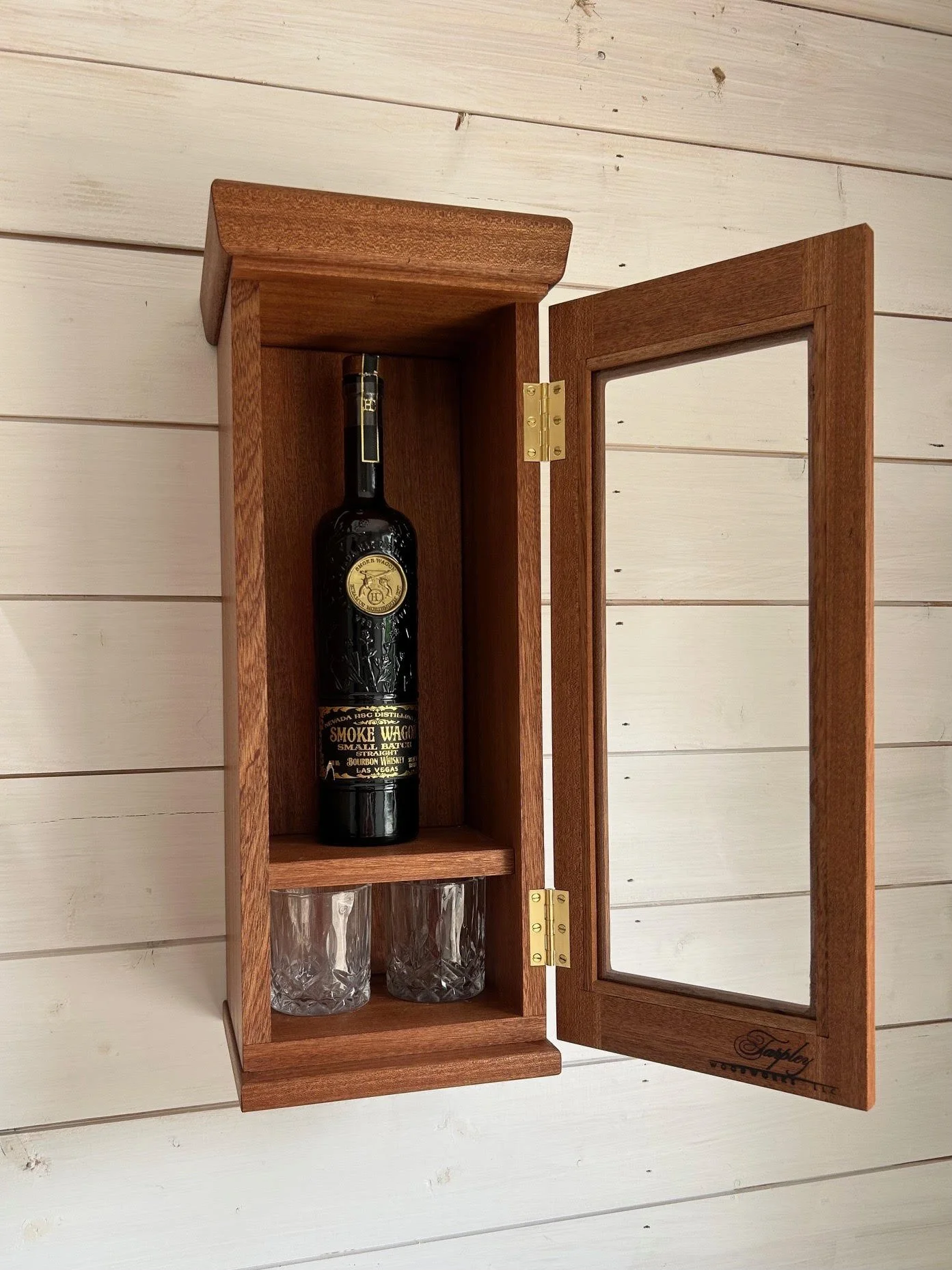 A wooden cabinet with a glass door, containing a bottle of bourbon whiskey and two empty glasses inside.